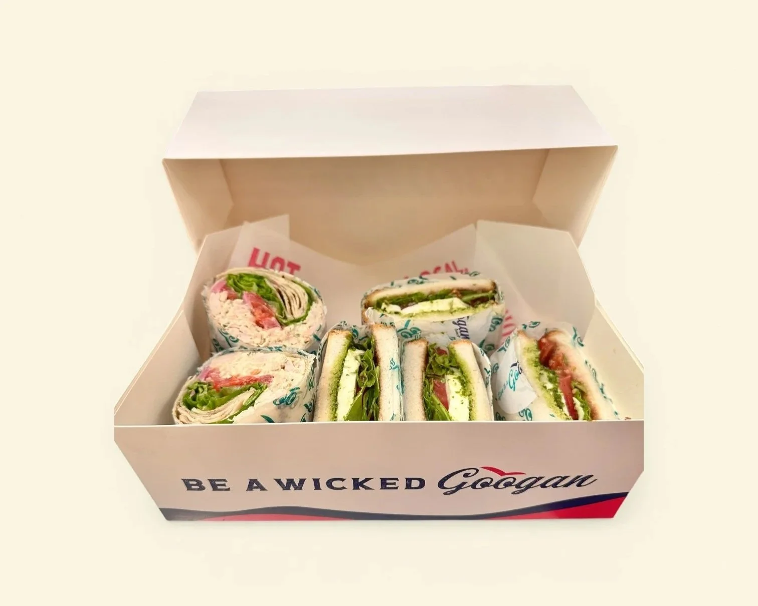 Open white box containing six wraps with lettuce, tomato, and other ingredients, lined with branded paper. The box has Red and blue text, including the phrase 'BE A WICKED Googan' on the side.