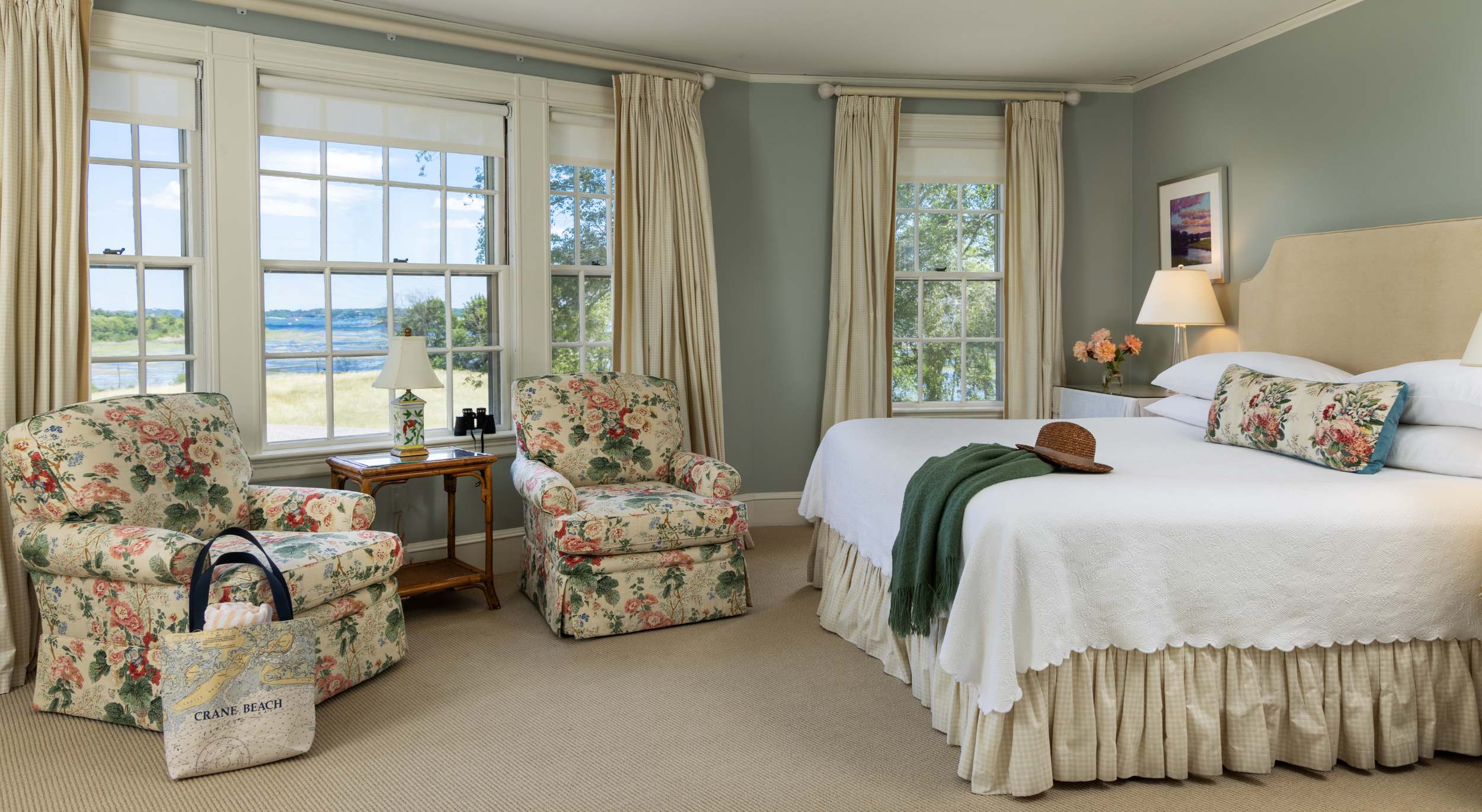 The Inn at Castle Hill. A cozy hotel room with a large bed, floral armchairs, and large windows showing a water view in an estate.