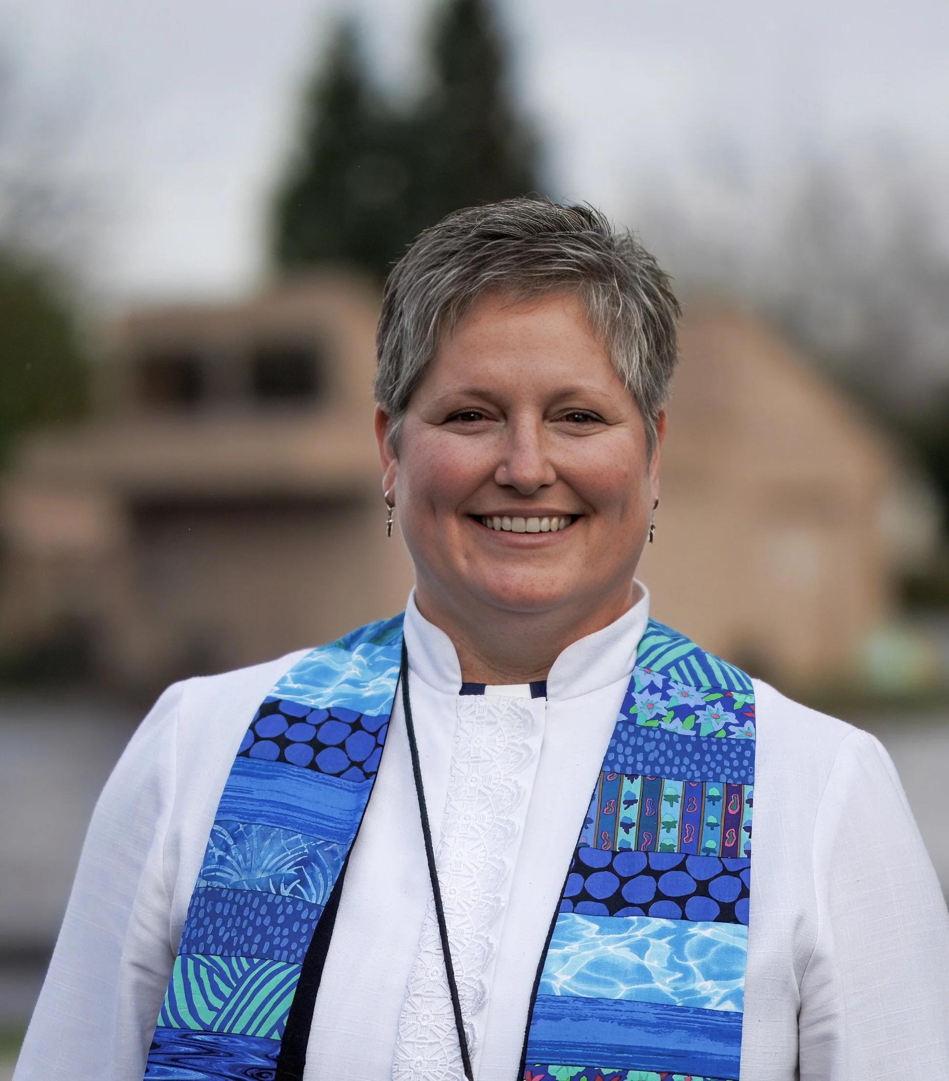 A woman with short blonde hair and glasses smiling outdoors with trees in the background.