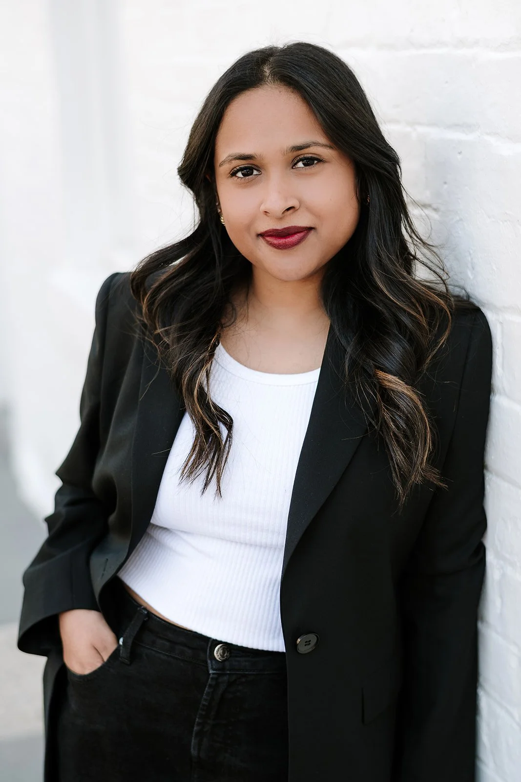 A woman with long dark wavy hair stands against a white brick wall, wearing a black blazer over a white top, with one hand in her pocket and a slight smile.