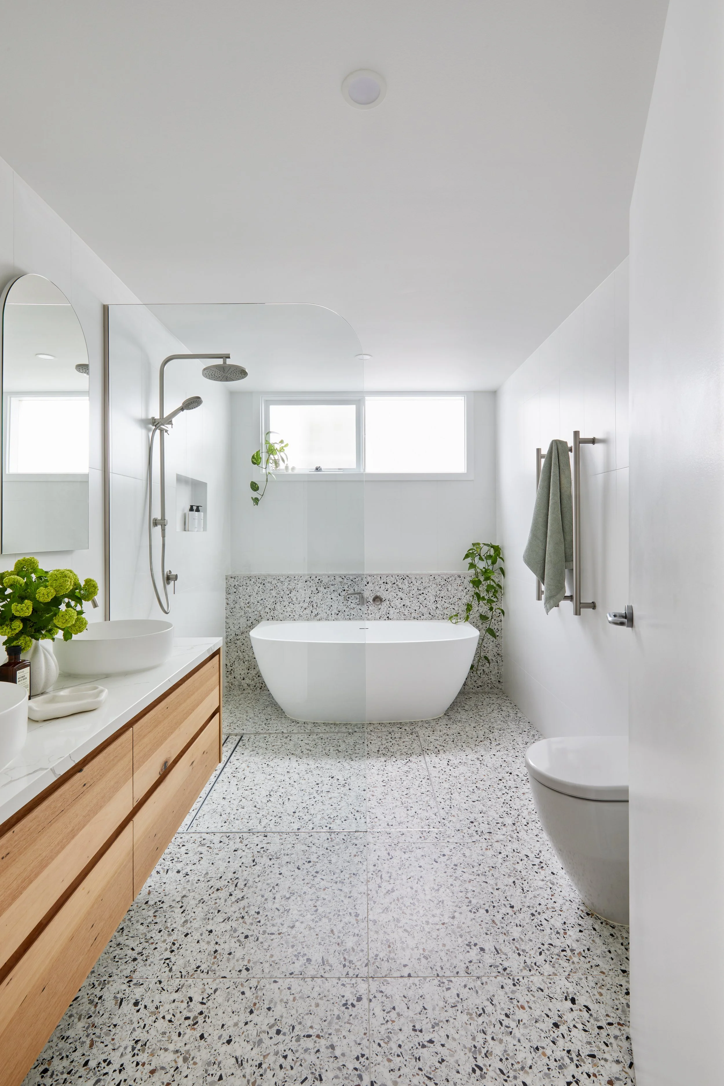 Modern bathroom with white walls and terrazzo flooring, featuring a freestanding bathtub near a small window, a wooden vanity with a vessel sink, a mirror, a shower with a rainfall showerhead, a wall-mounted toilet, and several green plants.