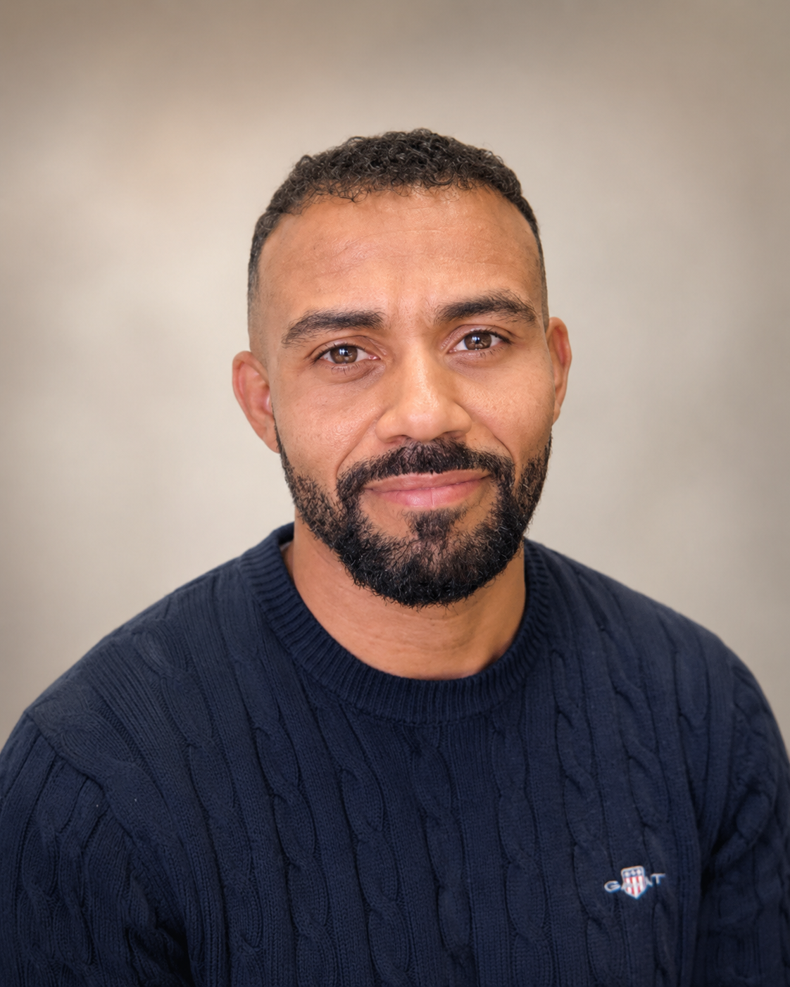 A man with short dark hair and a beard wearing a navy blue cable-knit sweater, sitting indoors in front of a plain wall.