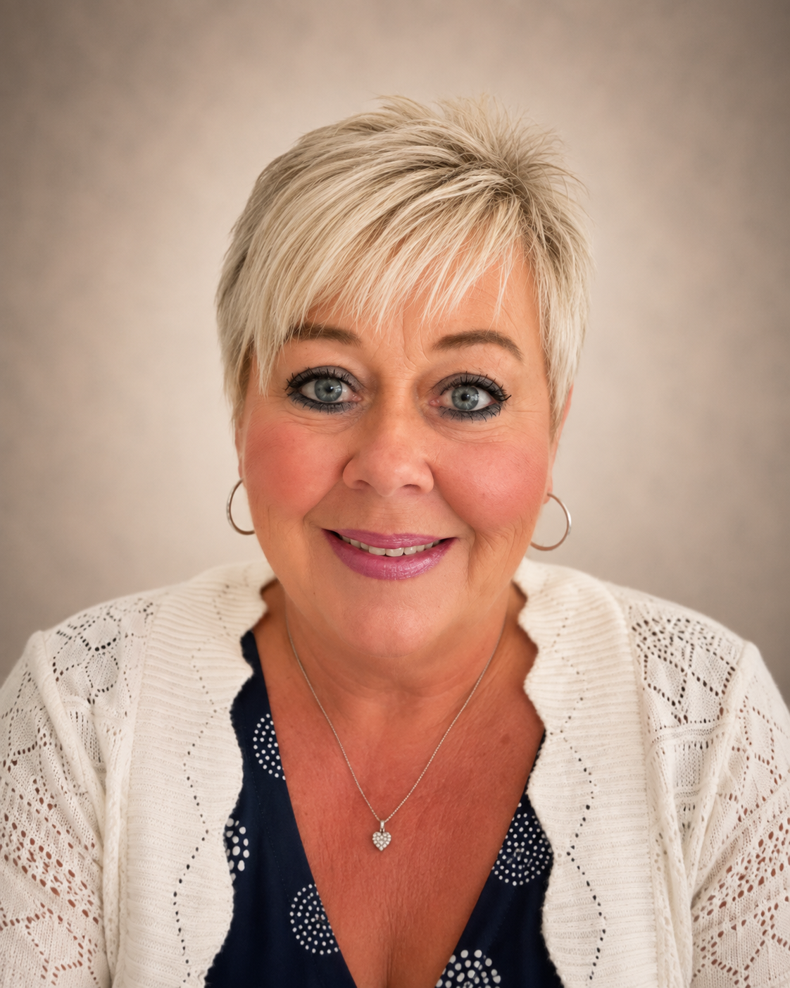 Close-up photo of a middle-aged woman with short blonde hair, blue eyes, wearing makeup, earrings, a necklace, and a cream-colored knitted cardigan over a black top with white polka dots, smiling in front of a neutral background.