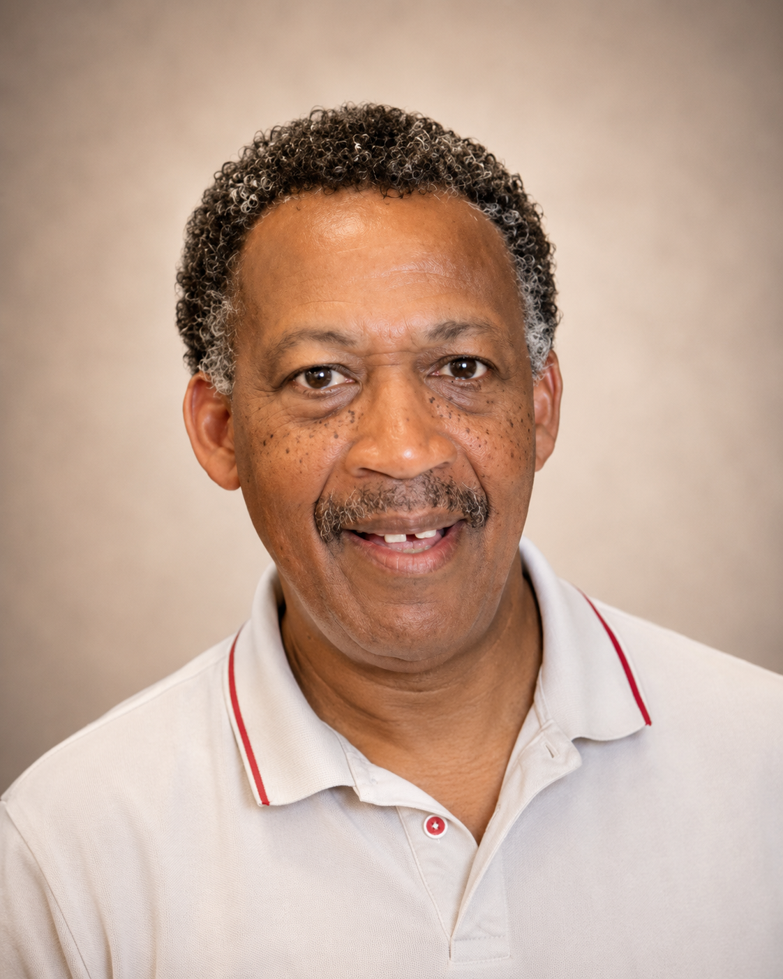 A smiling man with short curly hair and a mustache, wearing a white polo shirt with red trim, seated against a wooden wall.