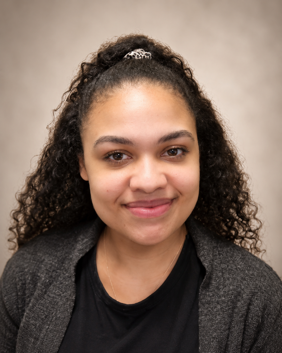 A young woman with curly hair pulled into a high ponytail with a scrunchie, smiling, wearing a black t-shirt that says 'JUST BE NICE' and a gray cardigan against a yellow background.