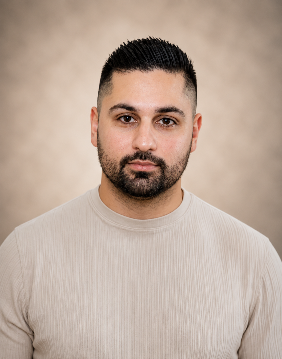 Portrait of a man with dark hair, beard, wearing a light-colored shirt, standing against a plain background.