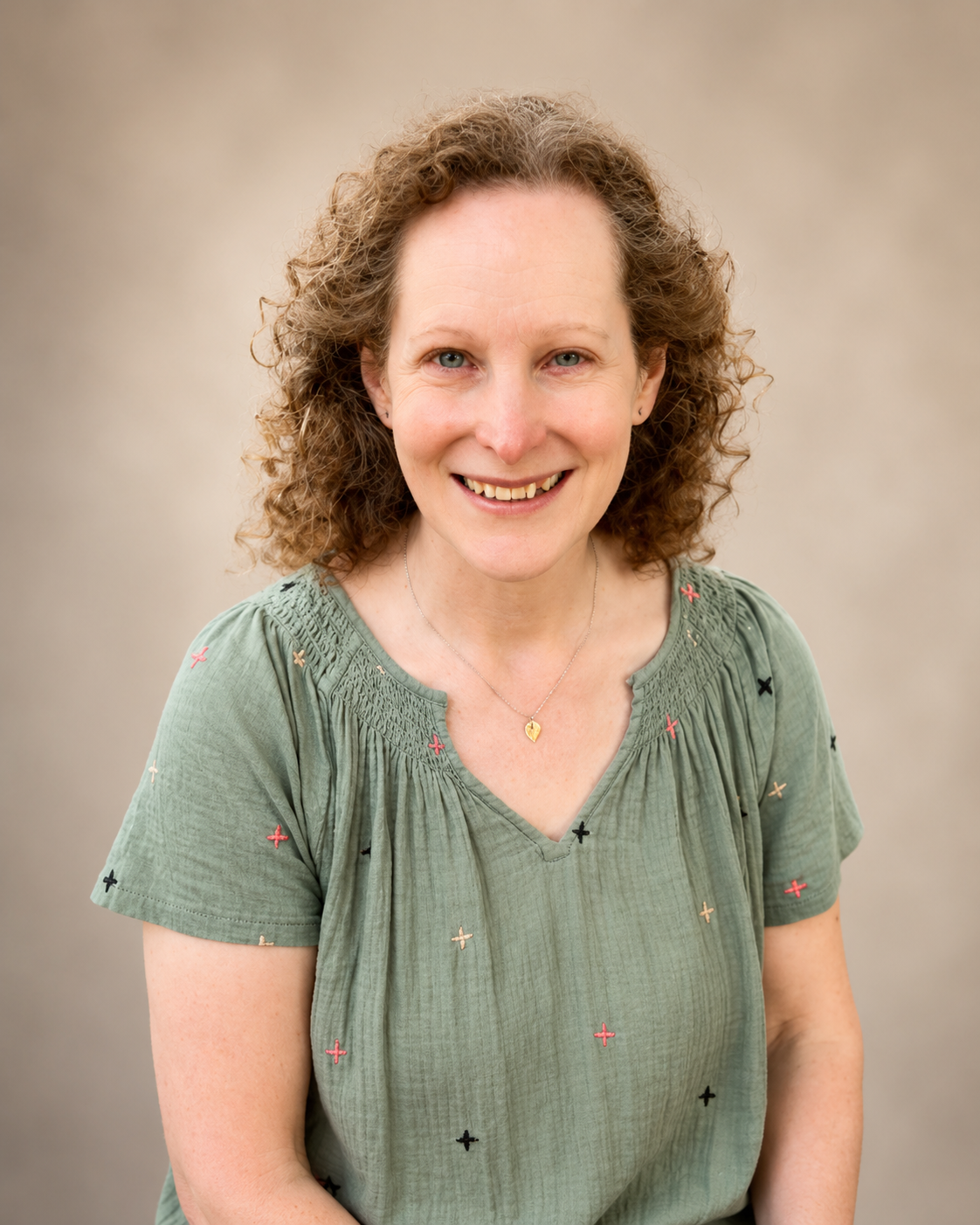 A woman with curly brown hair and blue eyes sitting on a wooden bench outdoors surrounded by green foliage and colorful flowers, smiling at the camera.