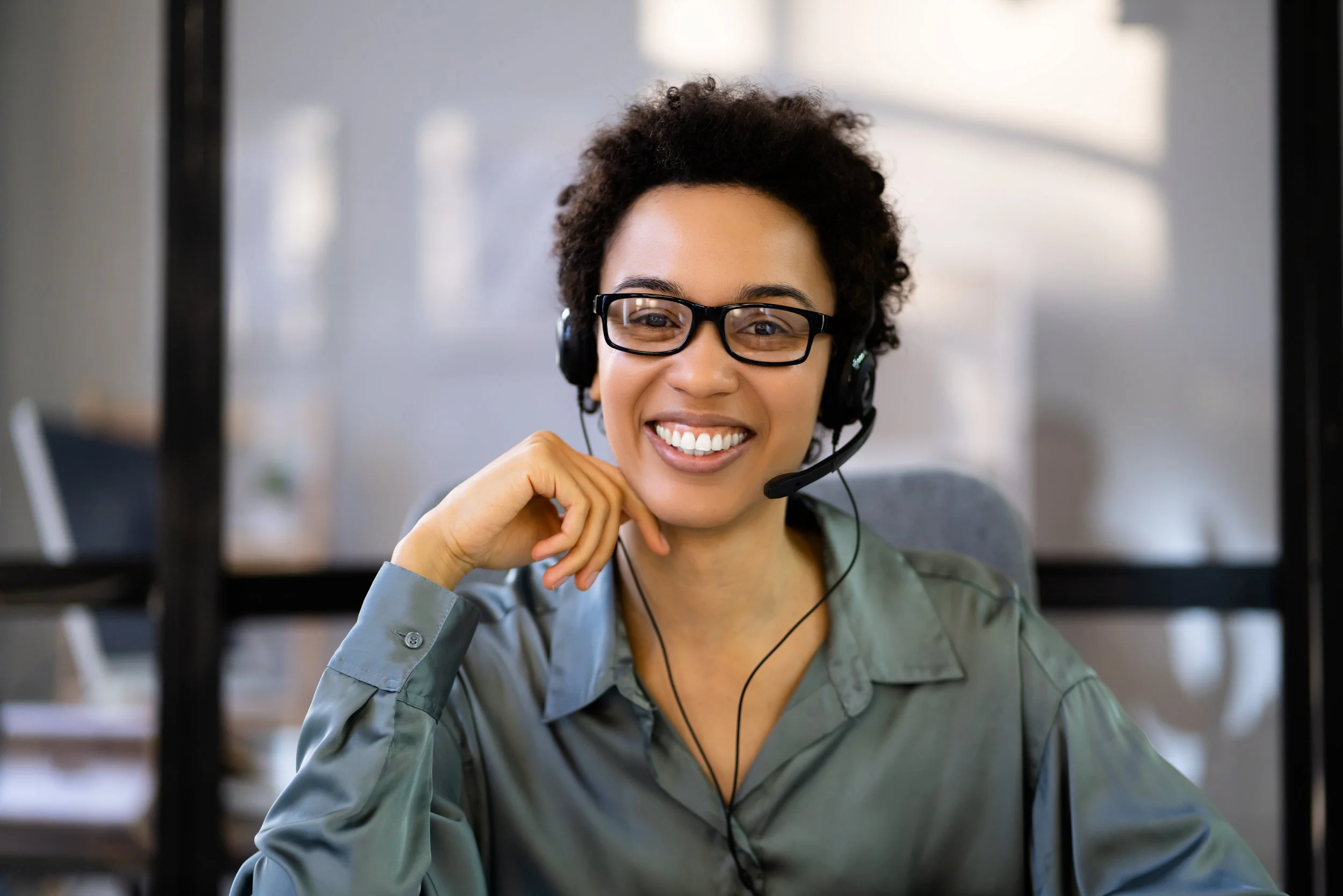 A woman wearing glasses and a headset, sitting at a desk and smiling.