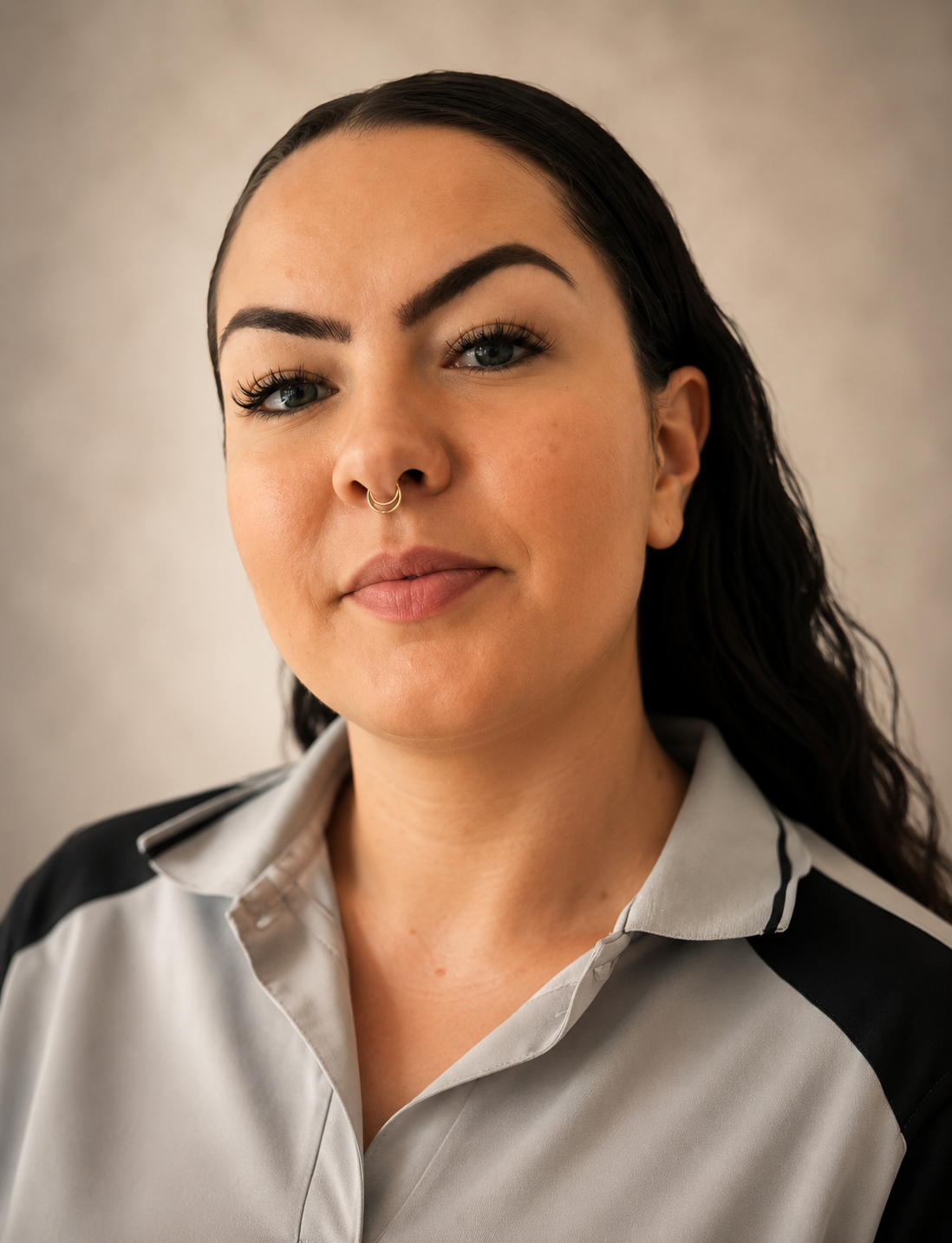 Close-up of a woman with dark hair, blue eyes, and a septum piercing, wearing a collared shirt, standing against a plain white wall.