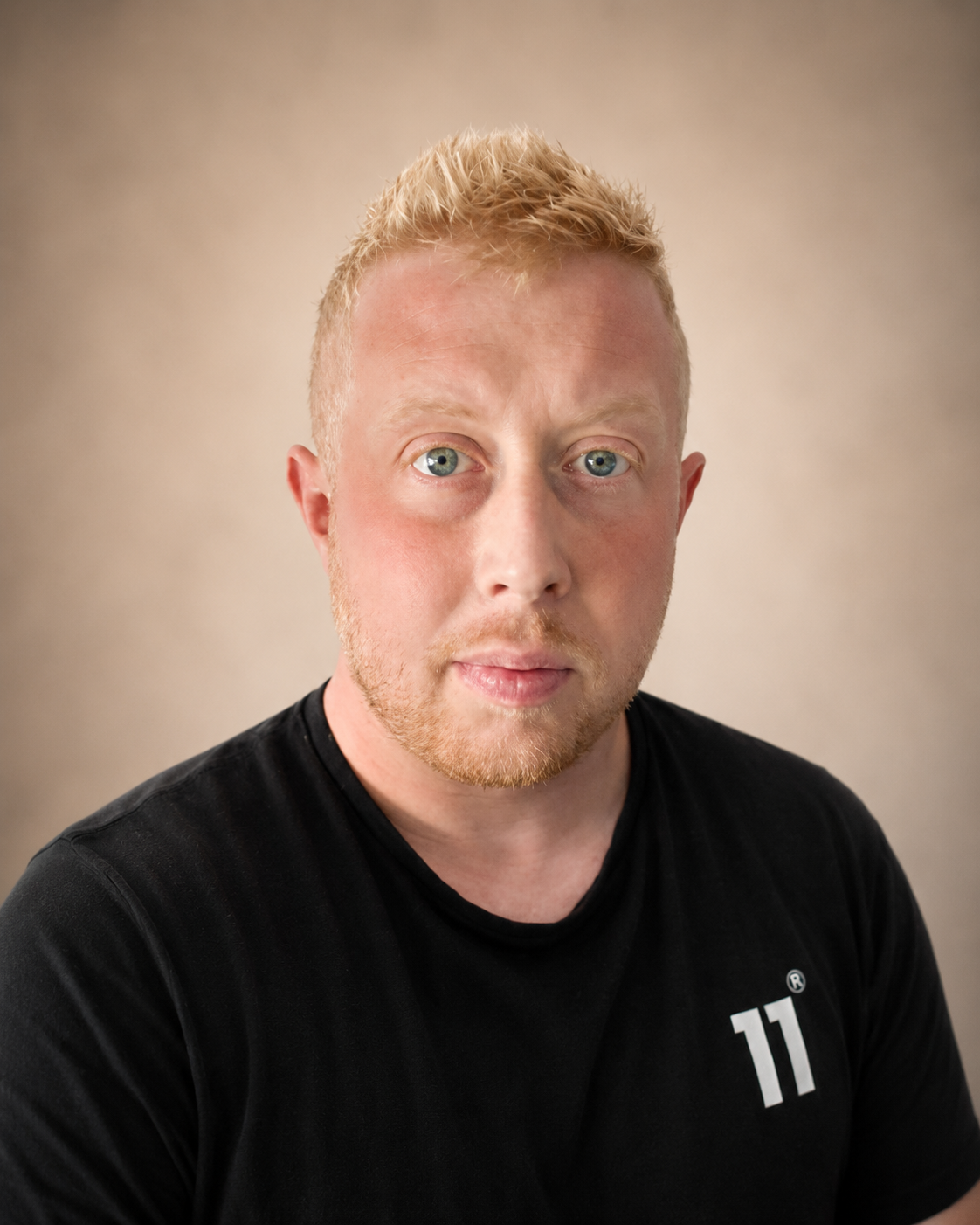 A man with ginger hair and blue eyes taking a selfie indoors, wearing a black shirt, with a blurred background that includes a mirror, a couch, and a window.