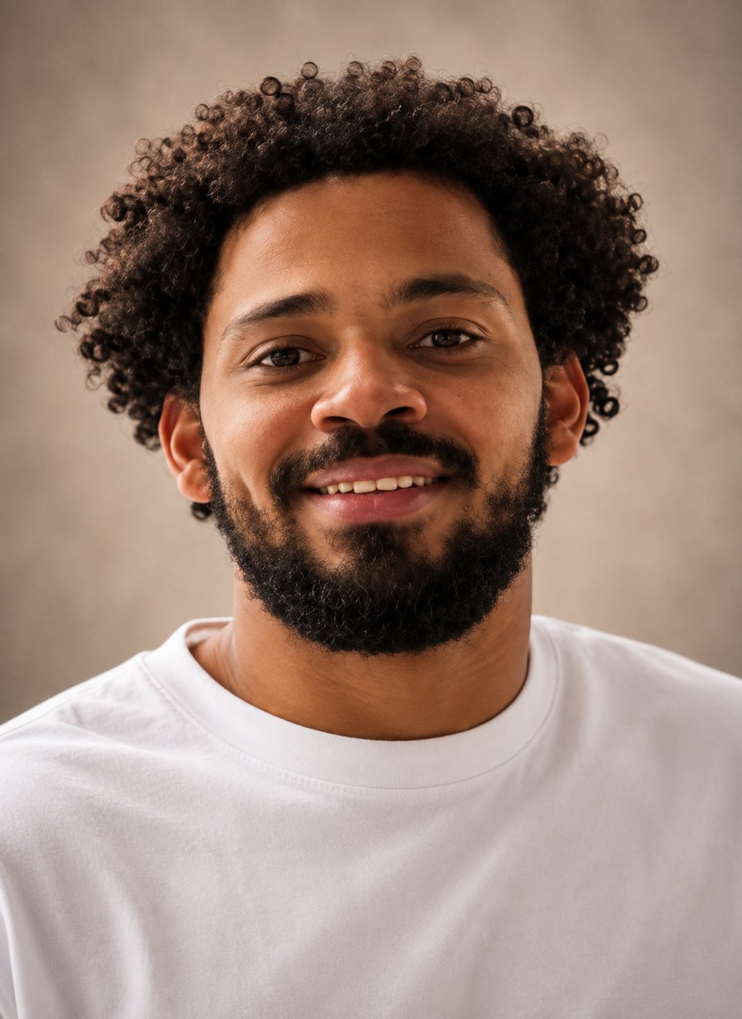 A young man with curly hair and a beard smiling at the camera, sitting indoors near a window.