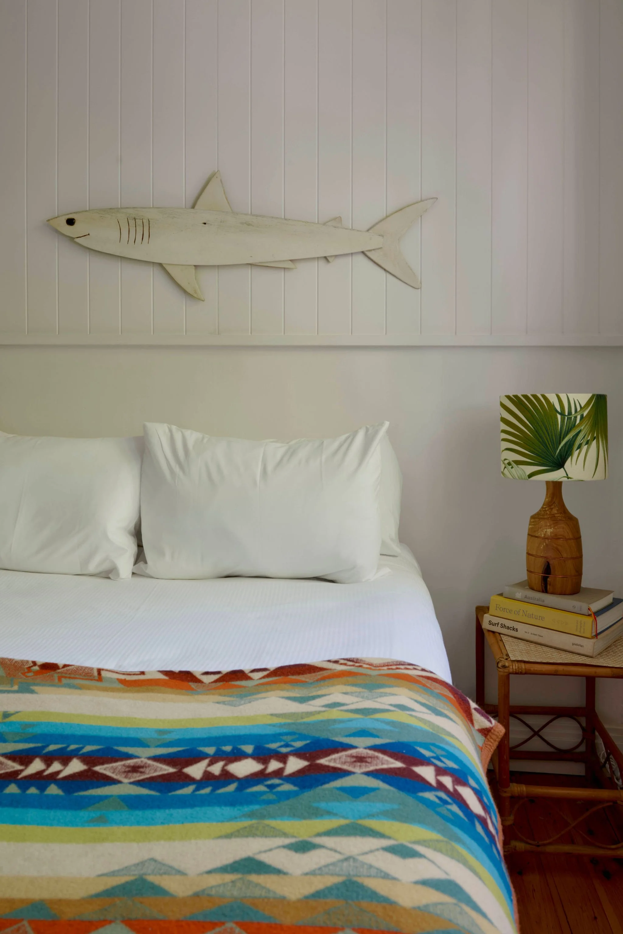 A bedroom featuring a wooden bed with striped and beige pillows, a white bedside table with a plant, and wall decorations including painted fish, with a blue door.