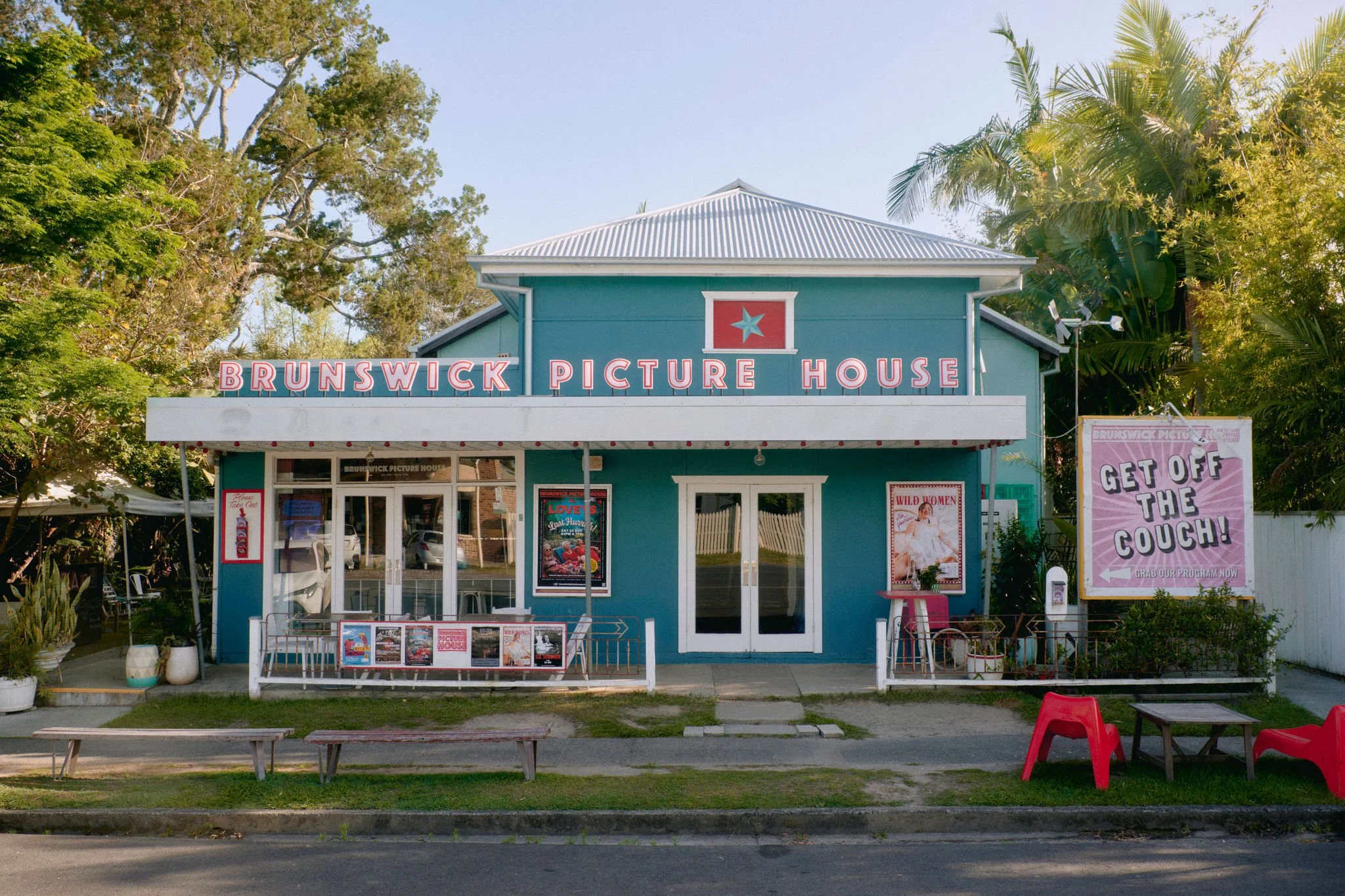 Night scene outside 'Brunswick Picture House'. People are gathered outside, some sitting, some standing and talking, with a bright billboard on the right saying 'Get Off The Couch'. The building has string lights and appears to be an entertainment venue.