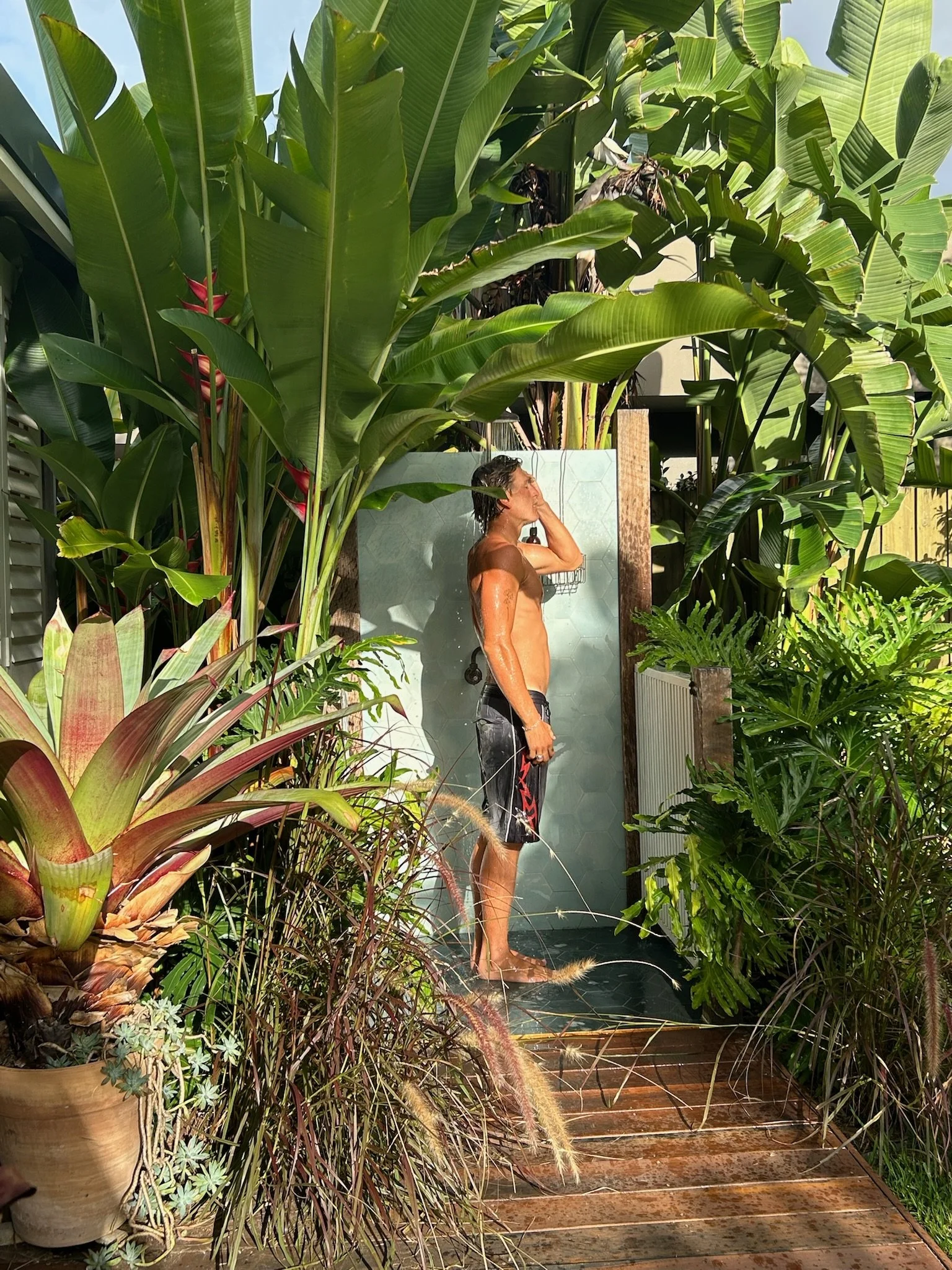 Outdoor shower with a yellow towel hanging on a light blue tiled wall and a wall-mounted basket holding a black soap dispenser, surrounded by lush green plants.