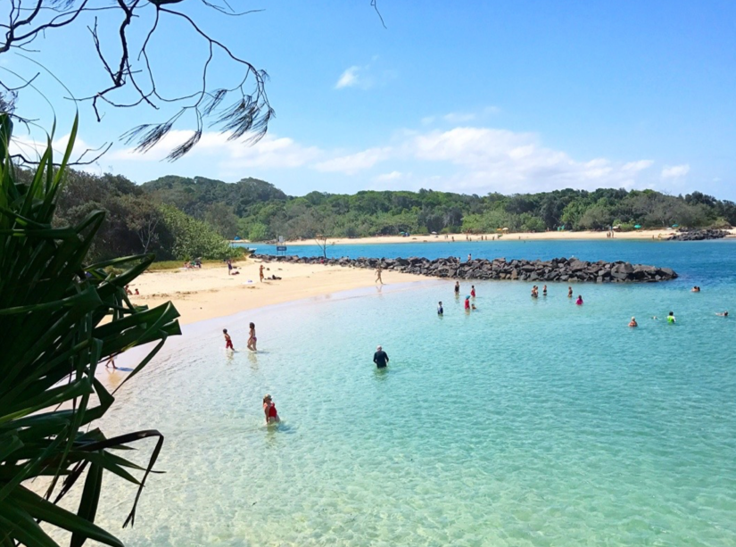 People swimming and relaxing at a clear blue beach with sandy shore, green trees, and a rocky breakwater under a partly cloudy sky.