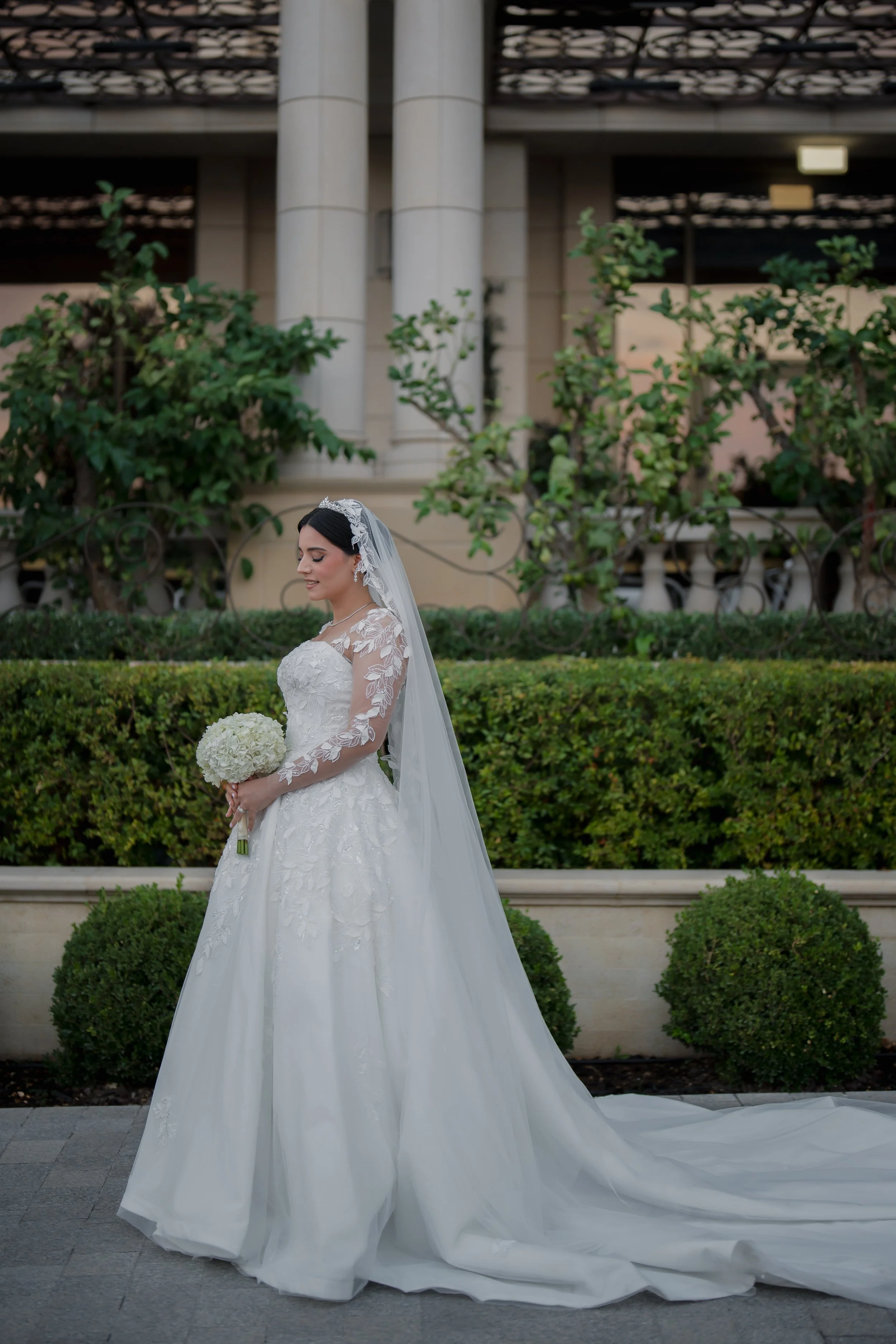 Bride in white wedding dress holding a bouquet, standing outdoors near greenery.