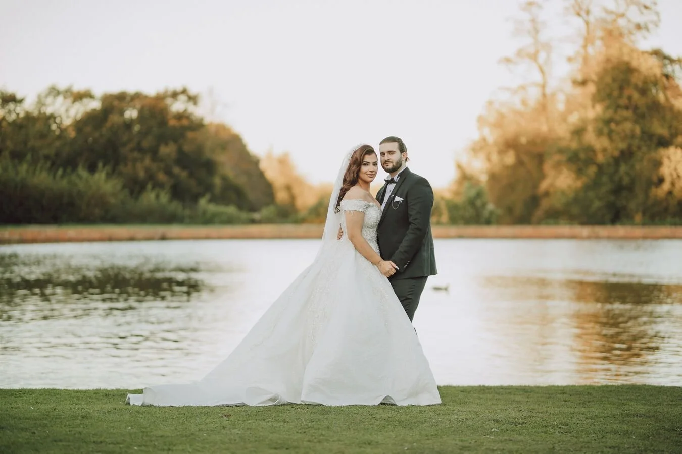 Bride and groom posing by a lake, outdoor wedding, sunset, elegant dress and tuxedo.