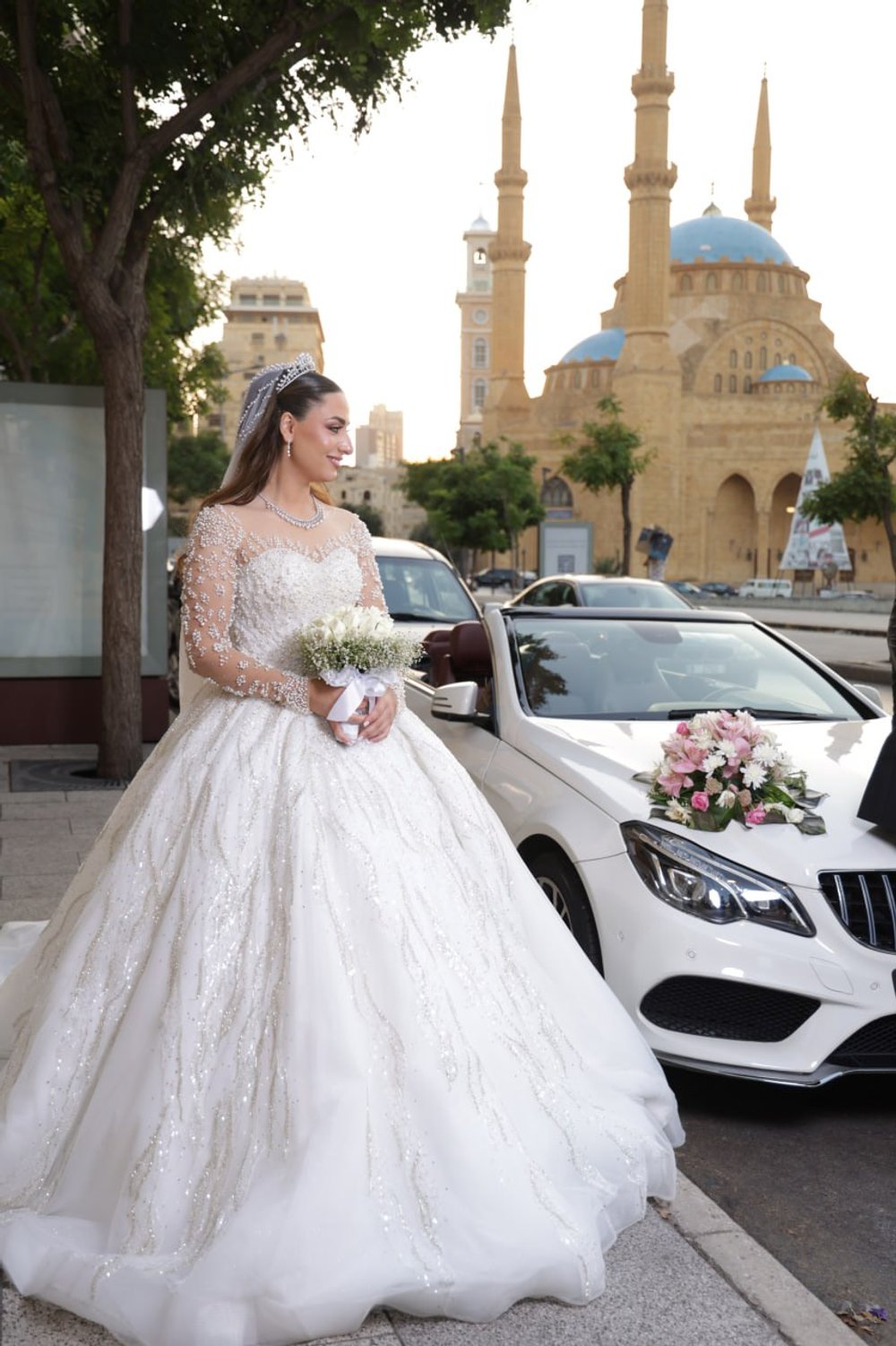 Bride in a white wedding gown holding a bouquet stands beside a decorated white car, with a mosque in the background.