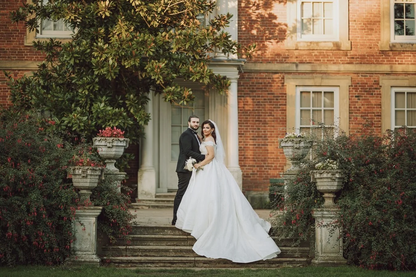 Bride and groom posing on steps in front of a brick building with greenery.
