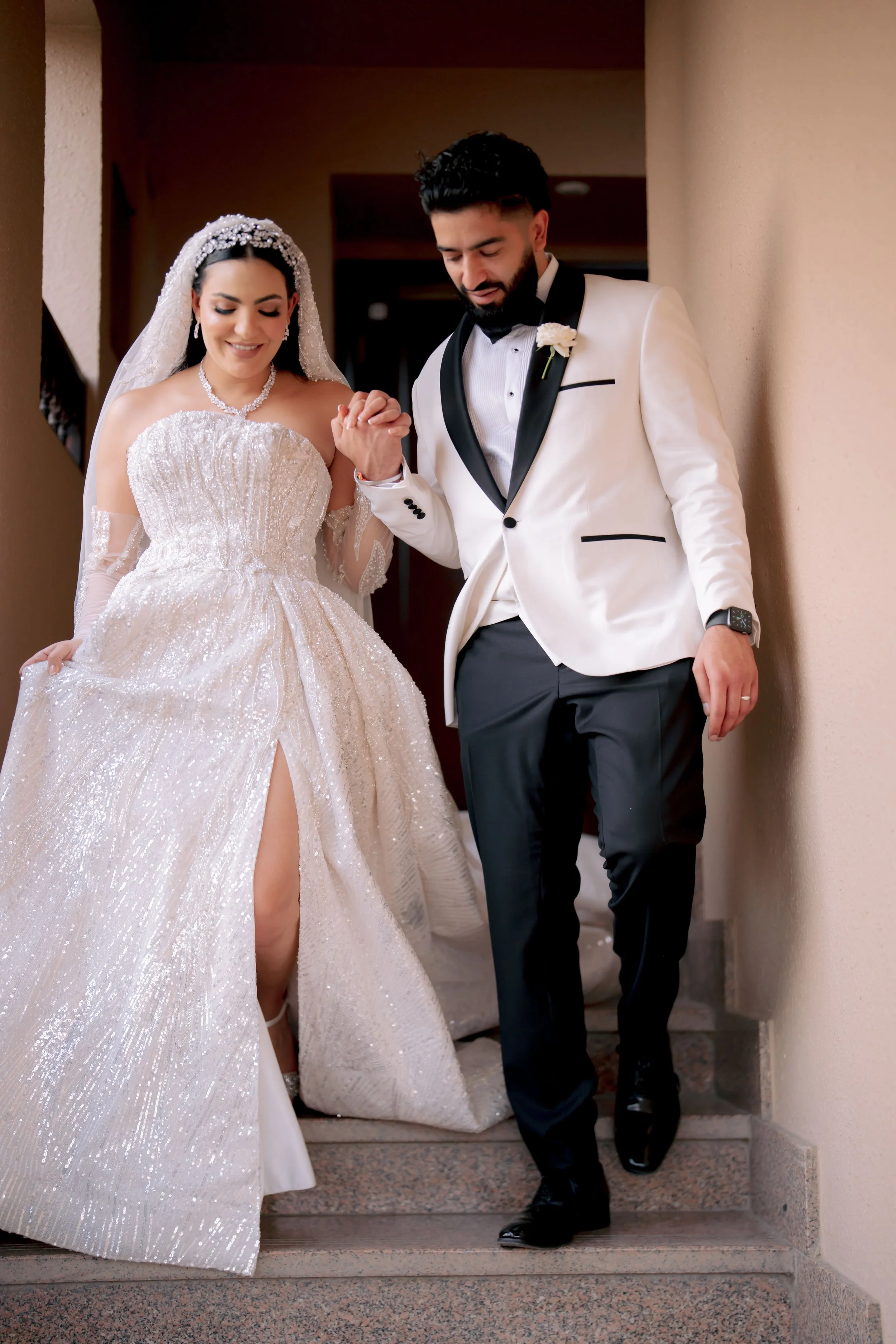 Bride and groom walking down stairs, bride in white gown, groom in white tuxedo with black accents.