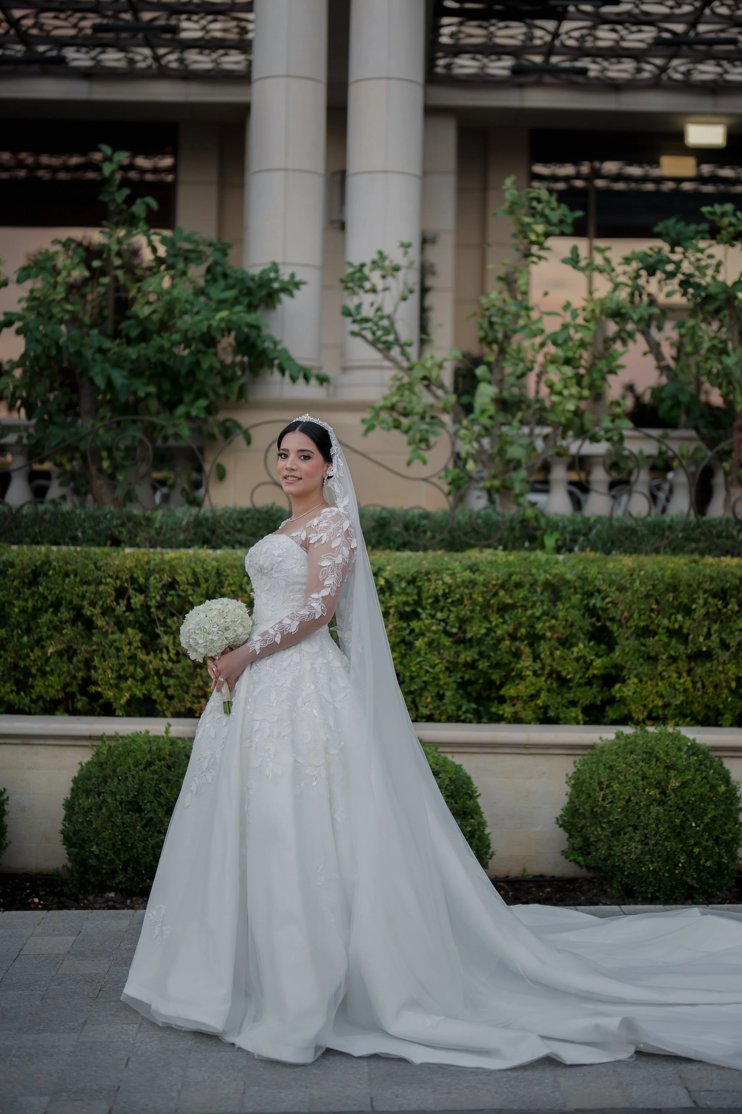 Bride in white gown holding a bouquet, standing outdoors with greenery and architectural columns in the background.