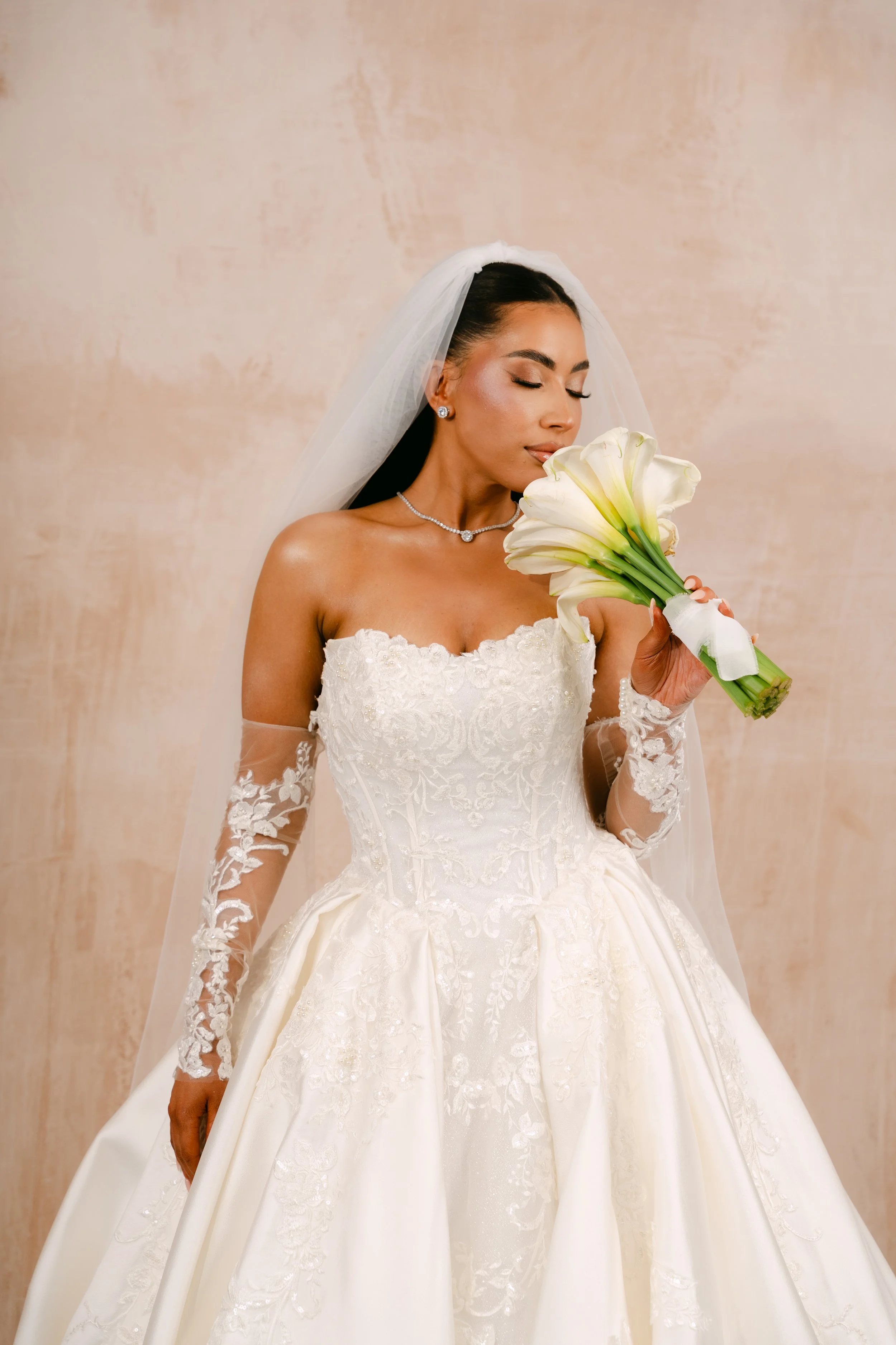 Bride in ornate wedding gown and delicate veil holding red roses on staircase.