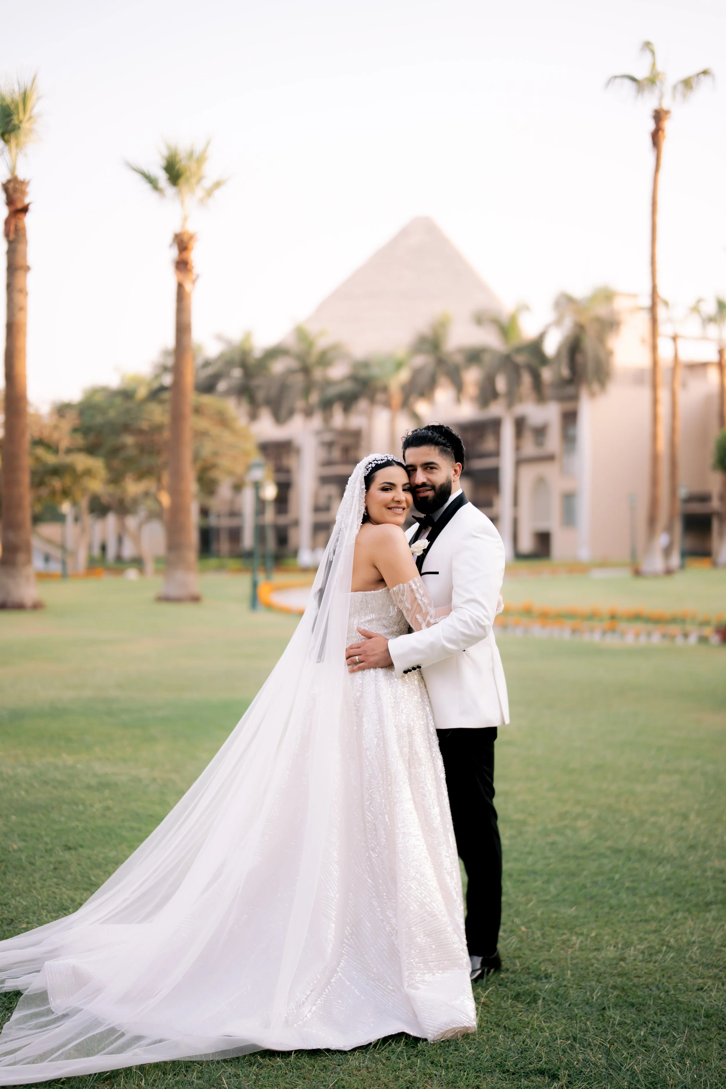 Bride and groom in wedding attire embracing in a garden with palm trees and a pyramid in the background.