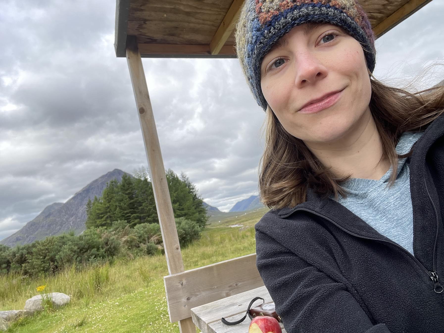 Carrie Dennis, writer, editor, content strategist, and artist wearing a beanie and sweatshirt, smiling for a selfie at a covered picnic table while outdoors on a cloudy day, in a nature setting with mountains, trees, and tall grass behind her