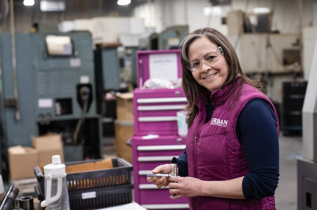 Woman in a manufacturing workshop wearing safety glasses and a purple Urban Manufacturing vest, standing near machinery and holding a tool.