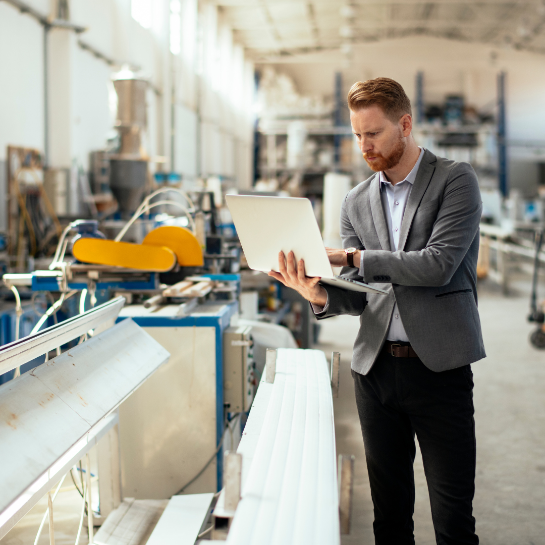 Business professional reviewing data on a laptop while standing inside a modern manufacturing facility, representing strategic communications and PR for manufacturing companies.