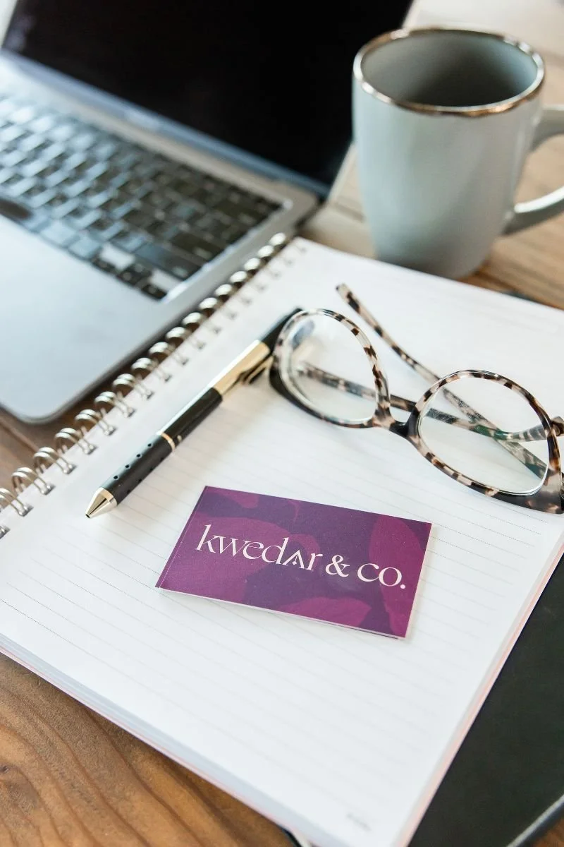 Kwedar & Co. business card with a notebook on a desk beside a laptop, pen, glasses, and coffee mug, representing thoughtful PR and marketing strategy work