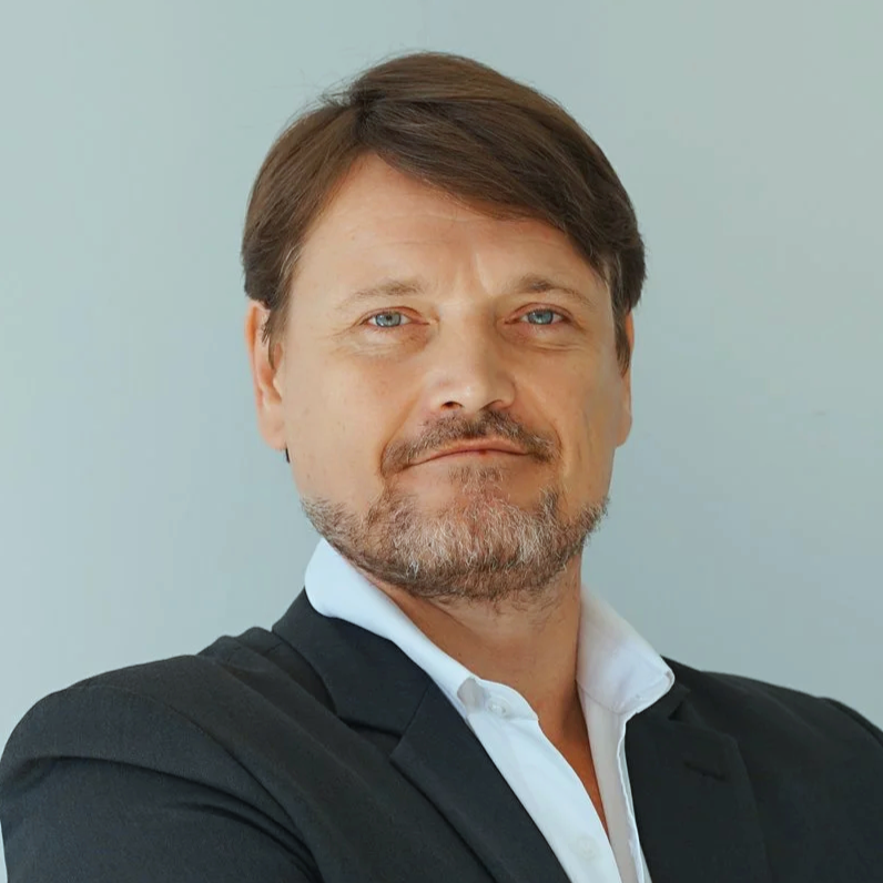 A confident man with light skin, brown hair, and blue eyes, wearing a black blazer and white shirt, posed with arms crossed against a plain, light background.