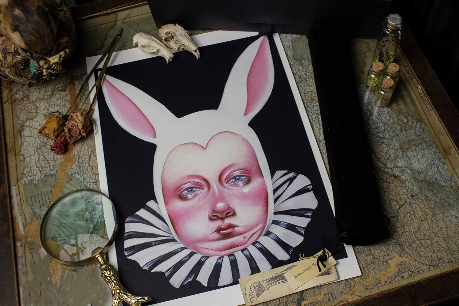 A surreal artwork of a human face with large eyes, pink cheeks, and lips, wearing a spooky white rabbit costume with long ears, set on a black background, surrounded by old dried flowers, a magnifying glass, and small glass bottles on a vintage map s