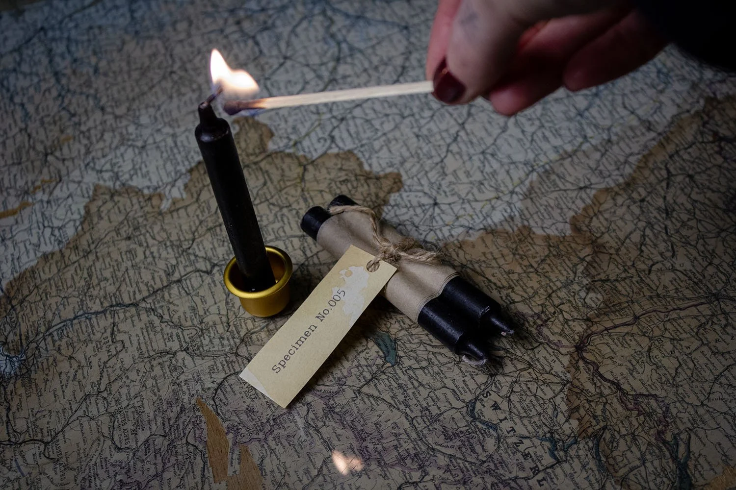 A matchstick is being struck on a black candle with a flame. The candle is part of a small rolled-up black object wrapped in brown paper, with a cream-colored tag labeled 'Specimen No. 005' tied to it. The items are placed on an old, torn map with va