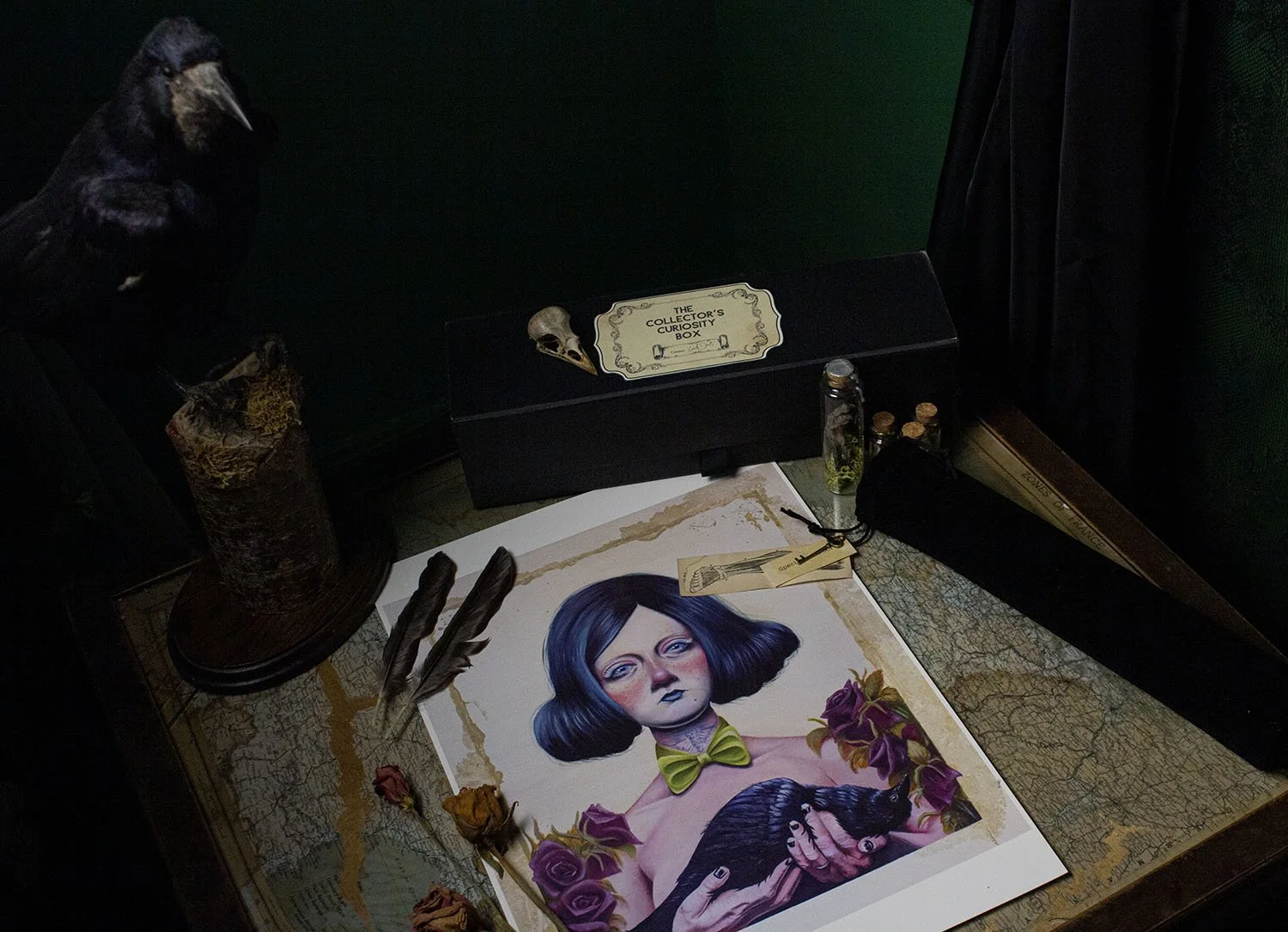 An art studio table with a detailed portrait of a woman with blue hair, purple flowers, and a bow tie, surrounded by dried roses and feathers. There is a black box labeled 'The Collector's Curiosity Box' and various small bottles nearby.