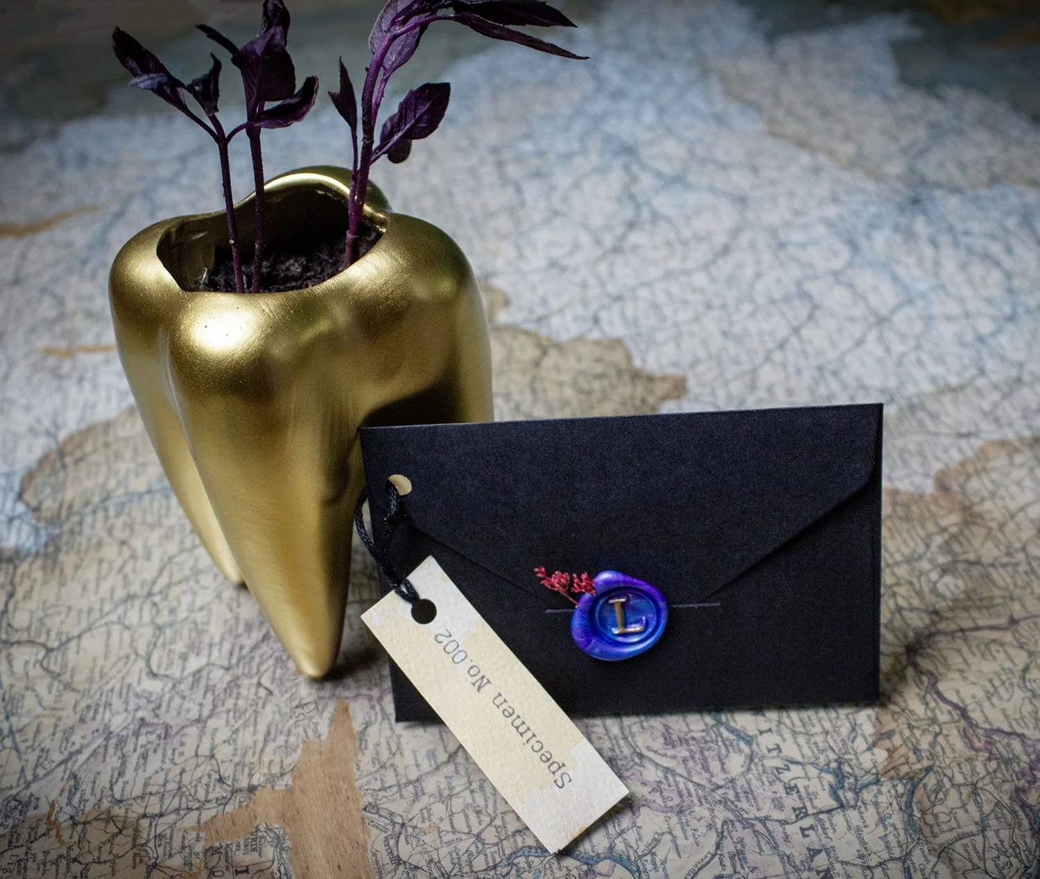 A gold-colored decorative tooth-shaped vase with purple plants inside, placed on a textured surface, next to a black envelope with a blue seal and a white tag labeled "Speeches No 200".