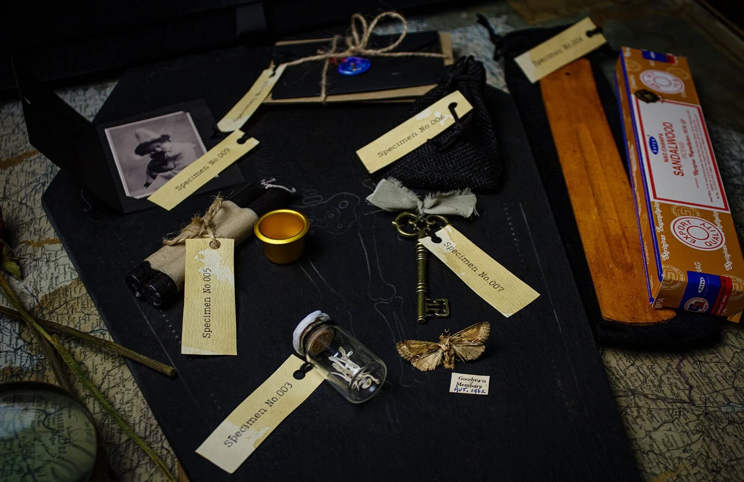 A collection of vintage and mysterious items, including an old contact's photograph, labeled specimens, a small vial with white objects, a butterfly, a key, a candle, and some mismatched items, laid out on a blackboard surface.