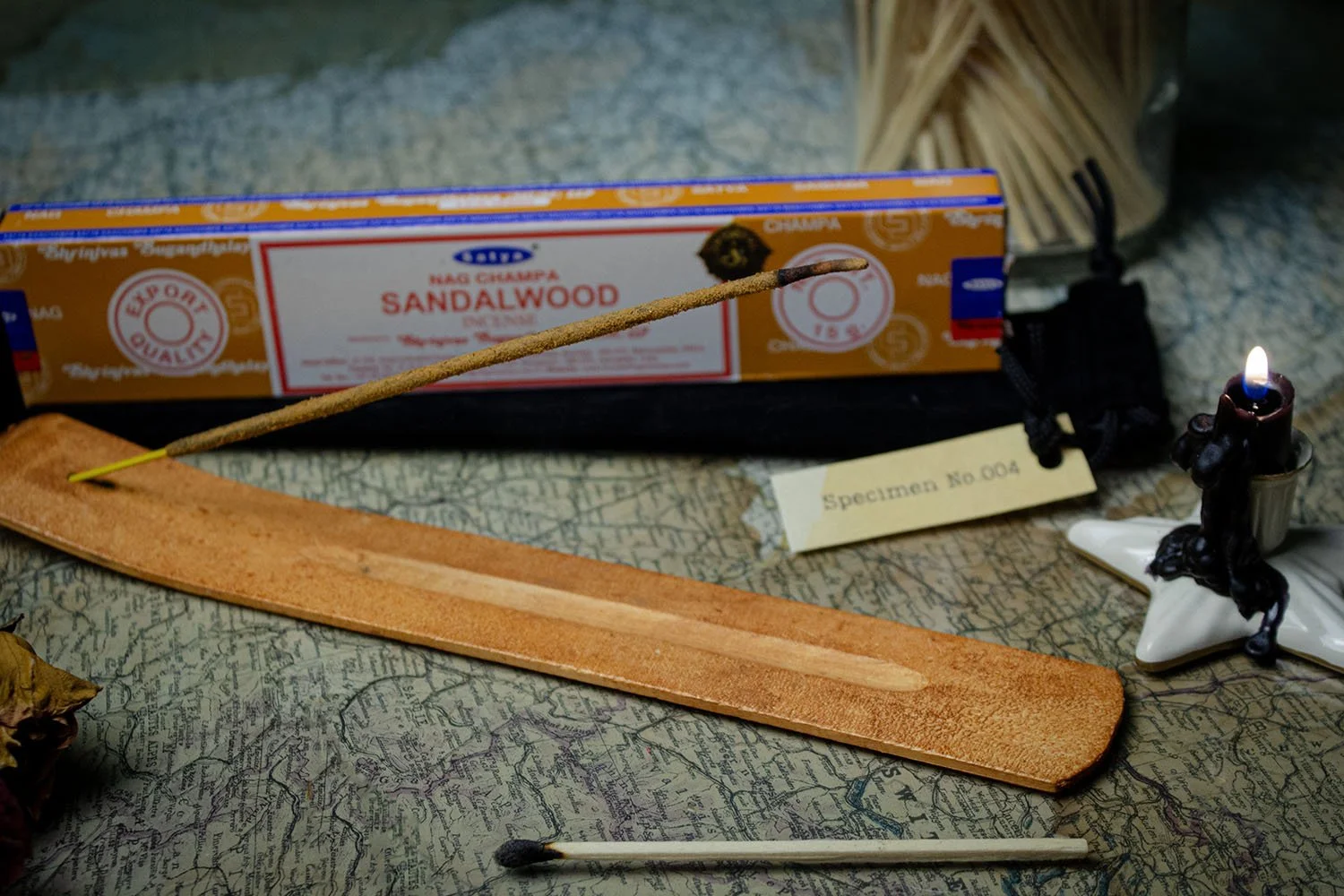 A matchstick with a burned tip placed on a wooden matchbook, with a box of Sandalwood incense sticks and a small candle holder with a figure lighting a candle in the background, arranged on a surface with a map pattern.