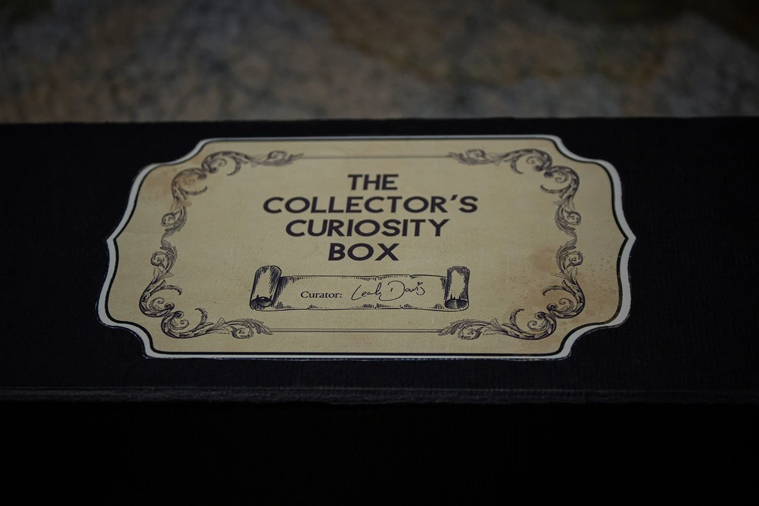 Close-up of a decorative label on a black box that reads "The Collector's Curiosity Box," curated by Leah Davis, with ornate borders and vintage style.