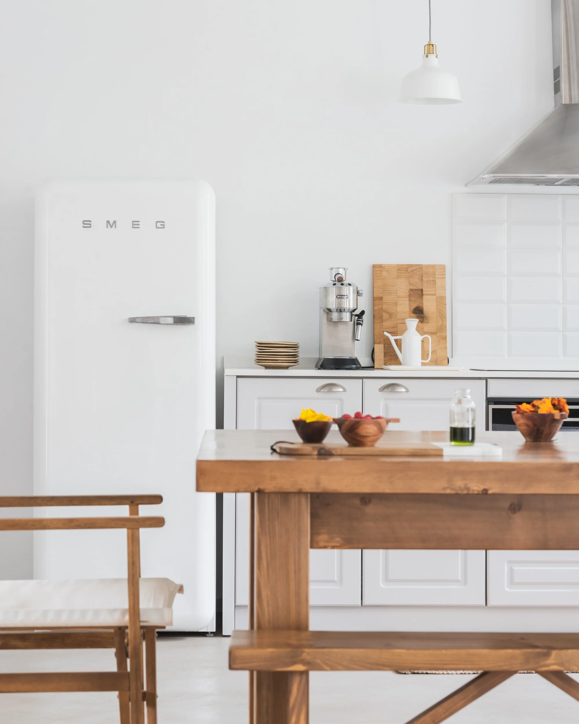 Cozinha moderna com geladeira branca SMEG, bancada branca, utensílios de cozinha de aço inox, painel de cortiça, prato de sobremesa, garrafa de azeite, conjunto de bowls de madeira, mesa de jantar e cadeiras de madeira