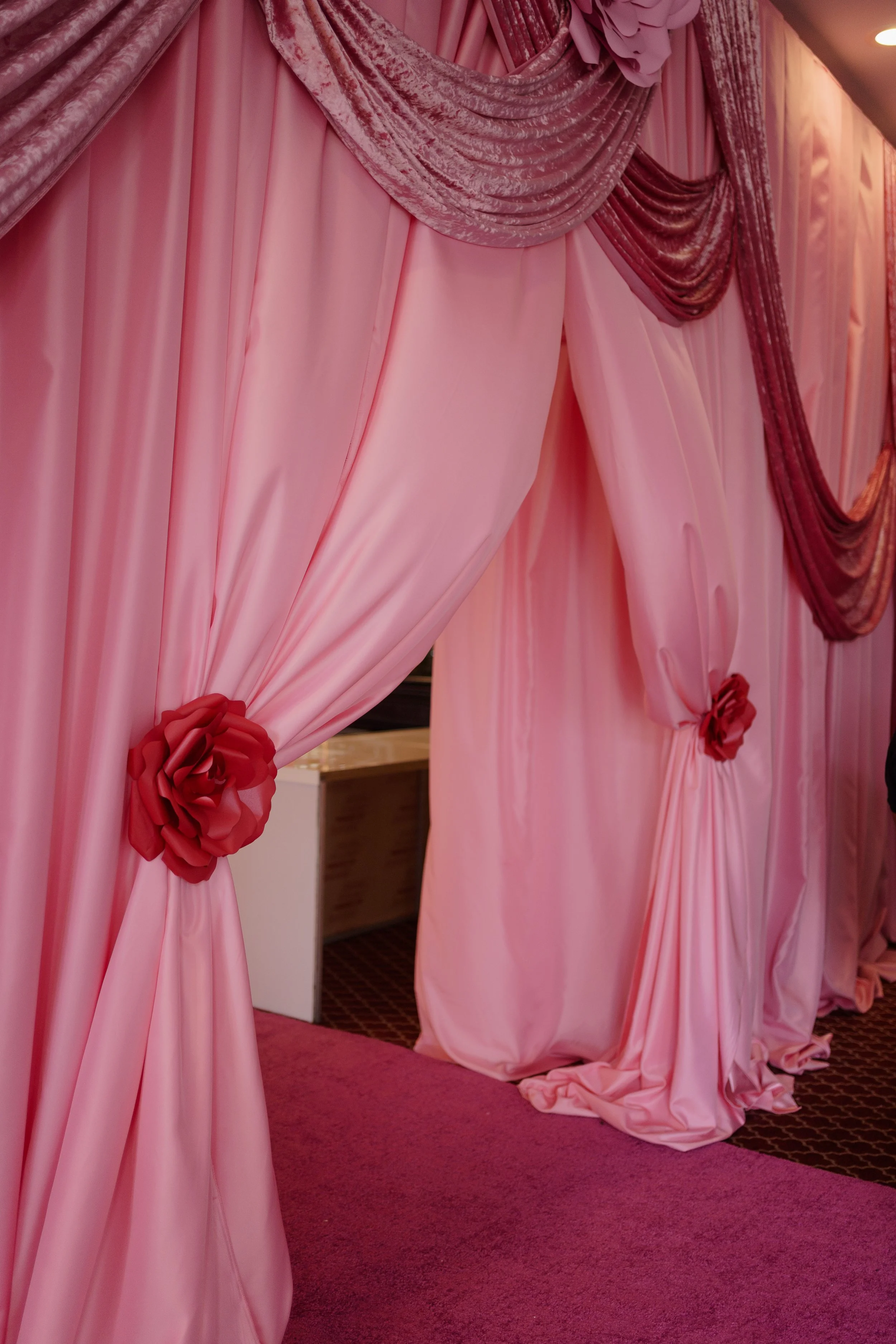 Pink velvet and satin curtain drapes with red fabric flowers as tiebacks and decorative swag at the top, set in an event space.