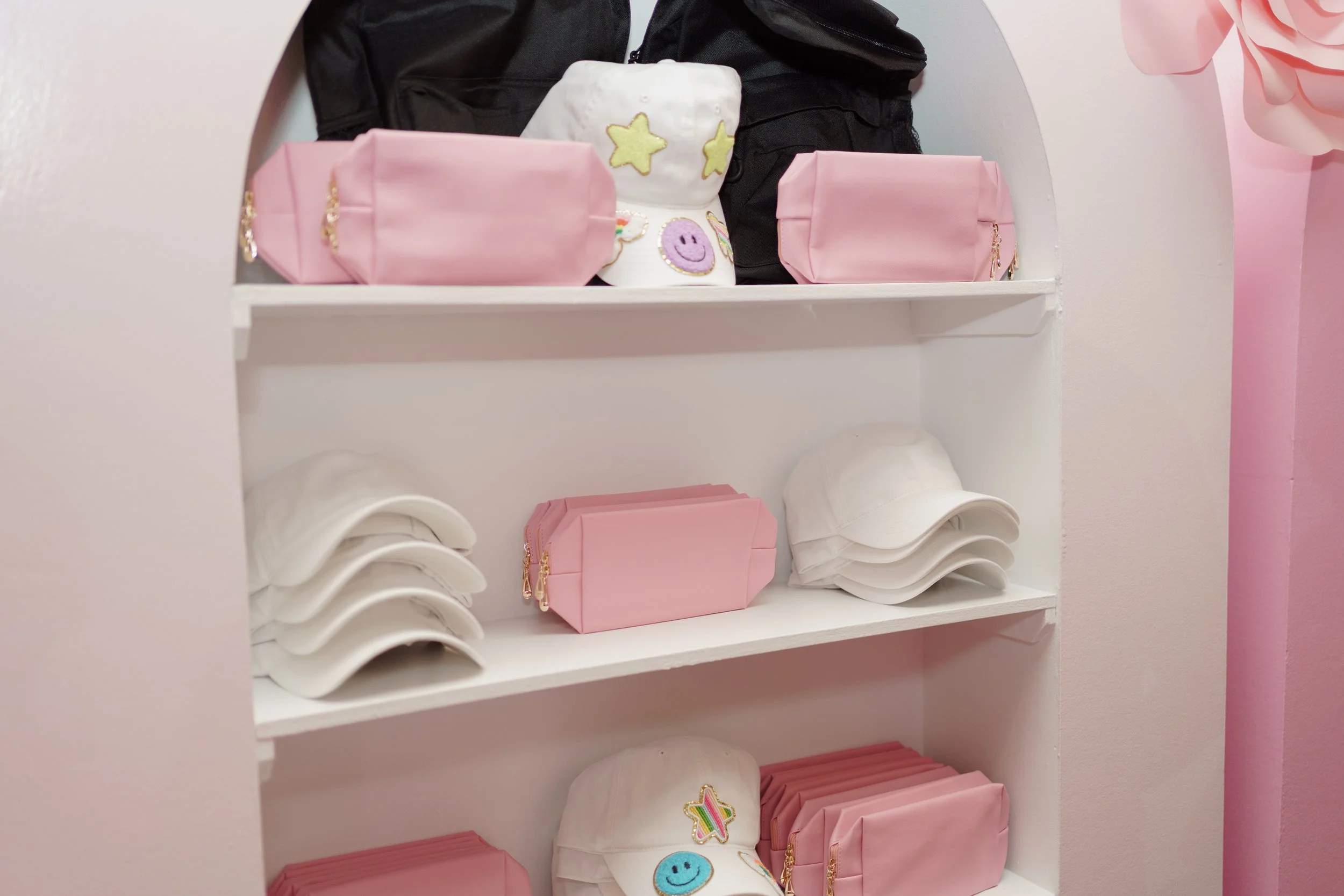White shelves holding pink and white caps with embroidered patches, pink cosmetic bags, white baseball caps, and black bags.
