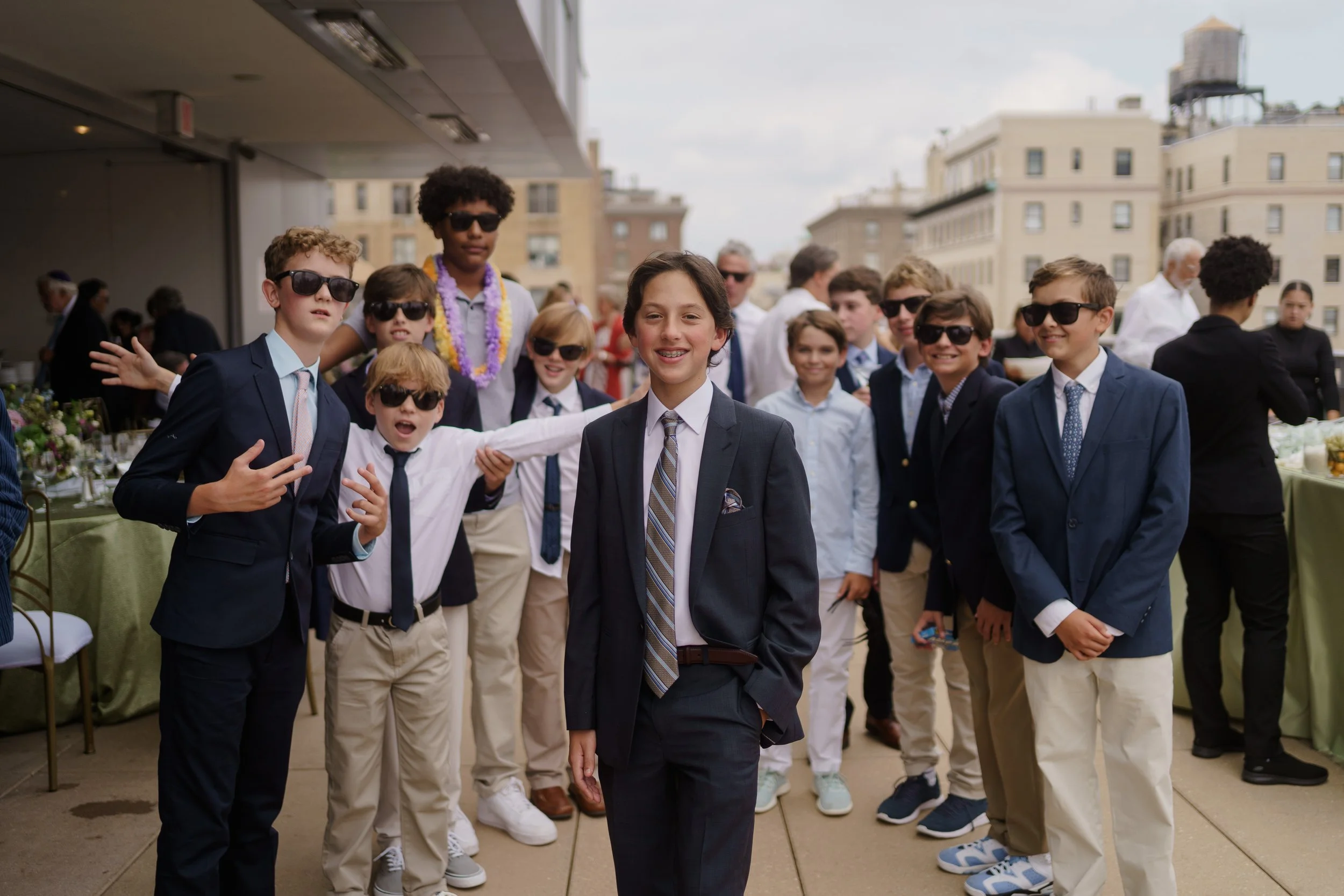 A group of boys and girls dressed in suits and casual clothing, some wearing sunglasses, at an outdoor event on a city rooftop with buildings in the background.