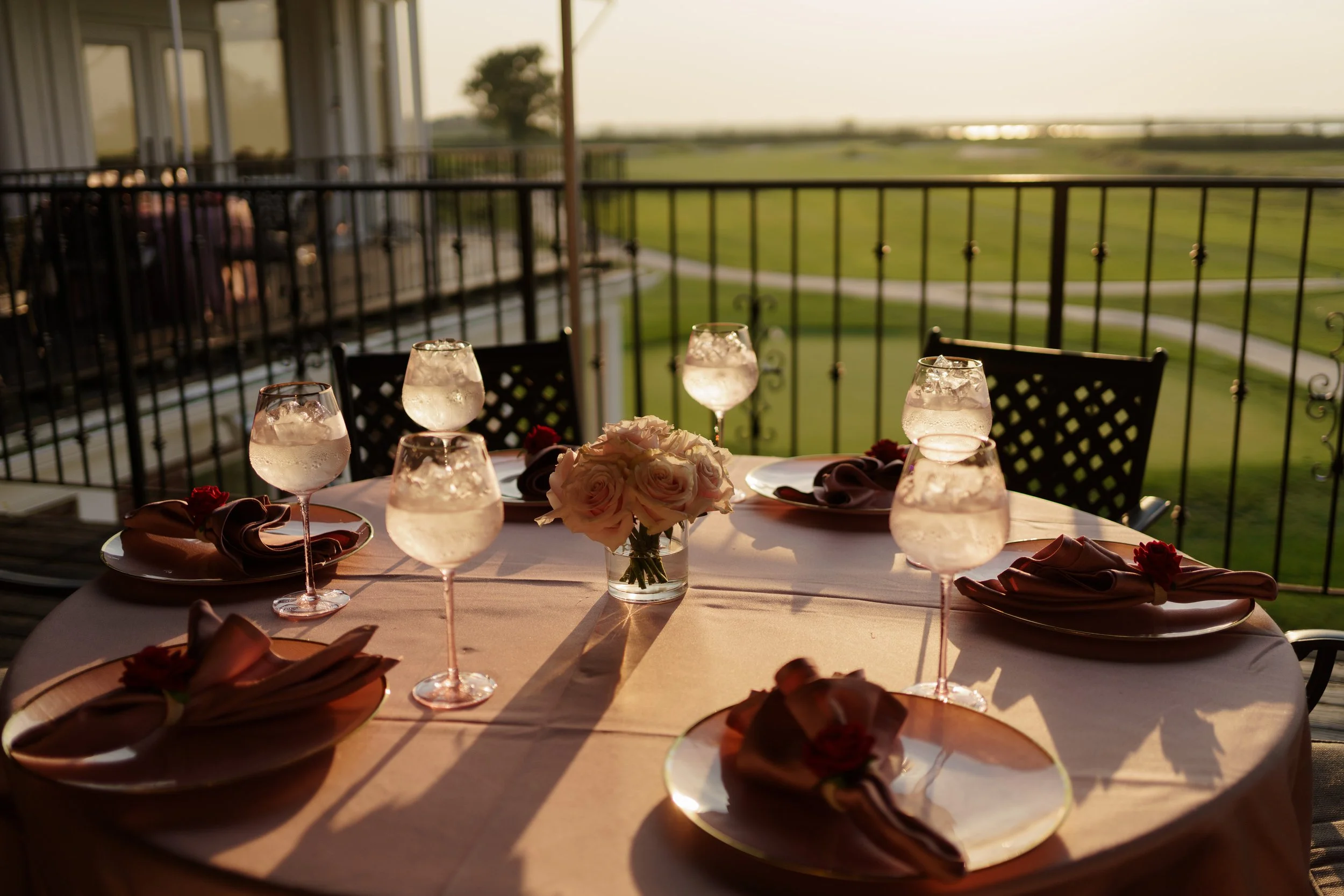 A round table set for a meal with white napkins, wine glasses with ice water, and a flower centerpiece, on a balcony overlooking a green landscape at sunset.