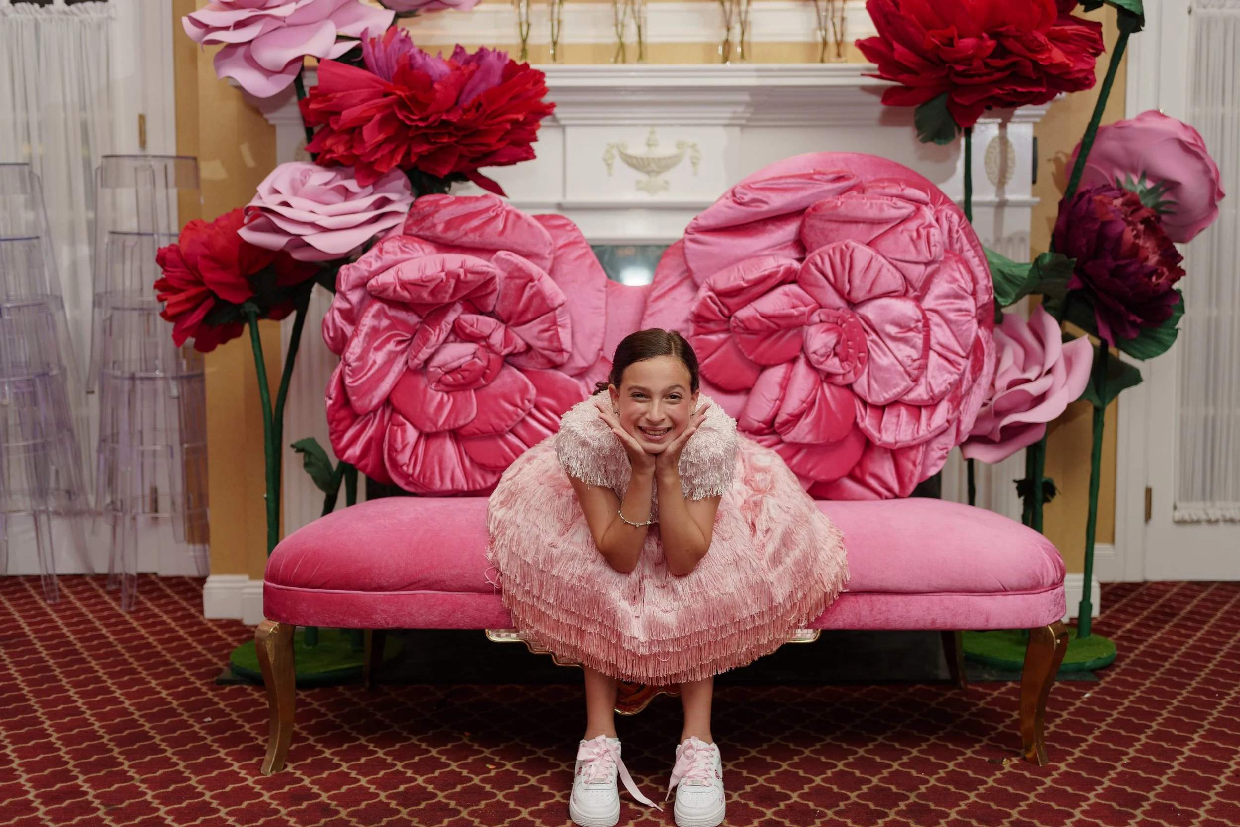 A young girl in a pink dress and sneakers is sitting on a pink velvet sofa with a large pink bow backrest, smiling and resting her chin on her hands, in a room decorated with giant pink and red paper flowers.