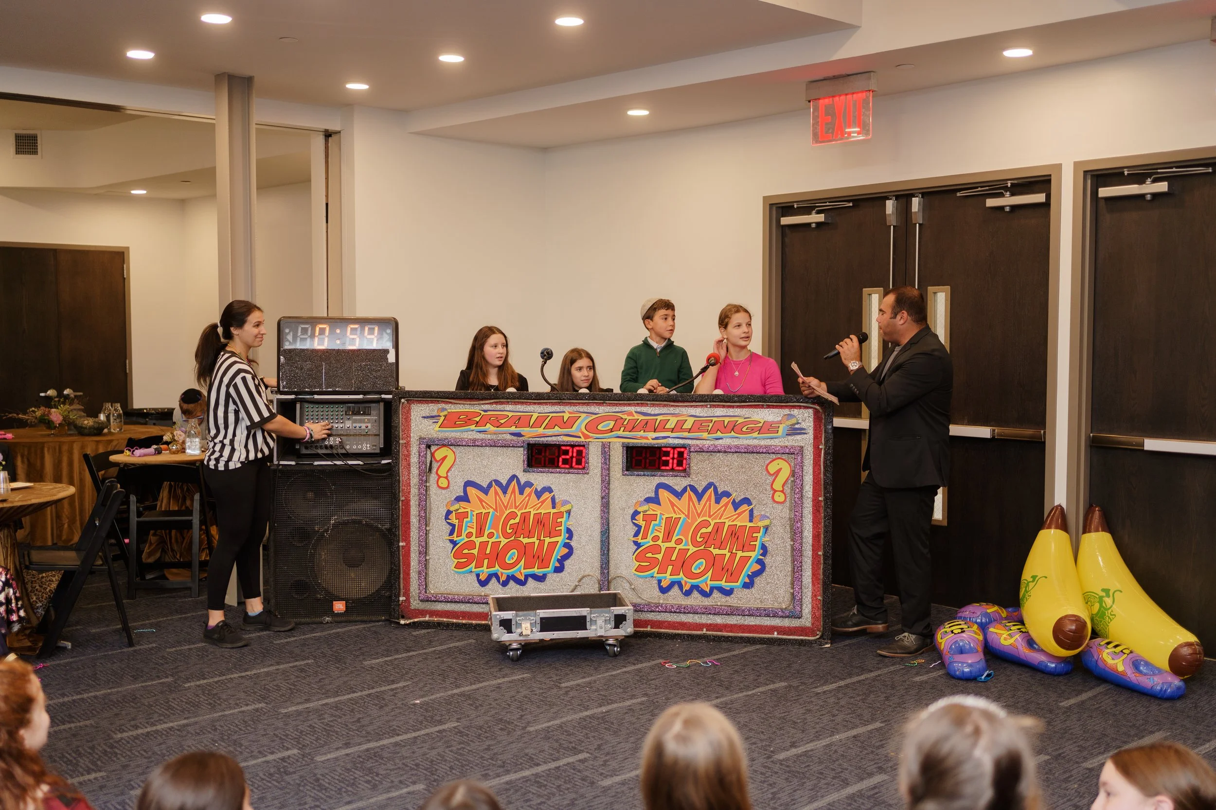 People participating in a game show called 'Brain Challenge' with a panel of four contestants, a host, and a referee, in a room decorated with inflatable bowling pins and shoes, with an audience watching.