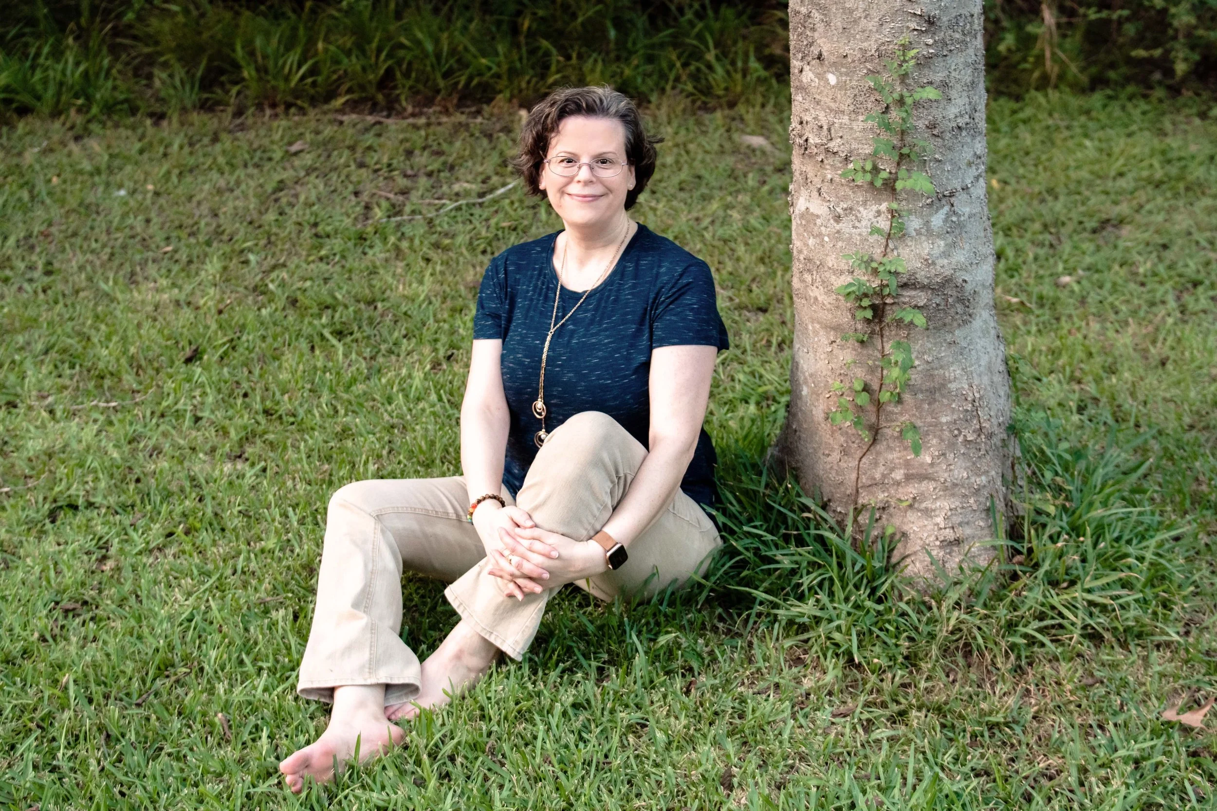 Dr. Christine Henry sitting on the grass by a tree crossed legged without shoes