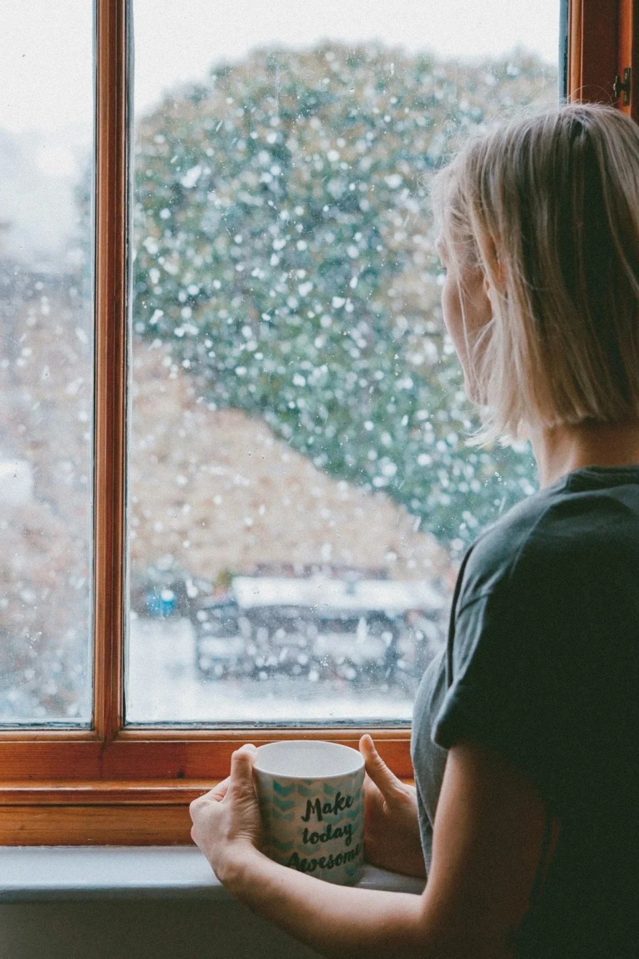 Woman standing next to a window with a mug to get time to decompress after a funeral