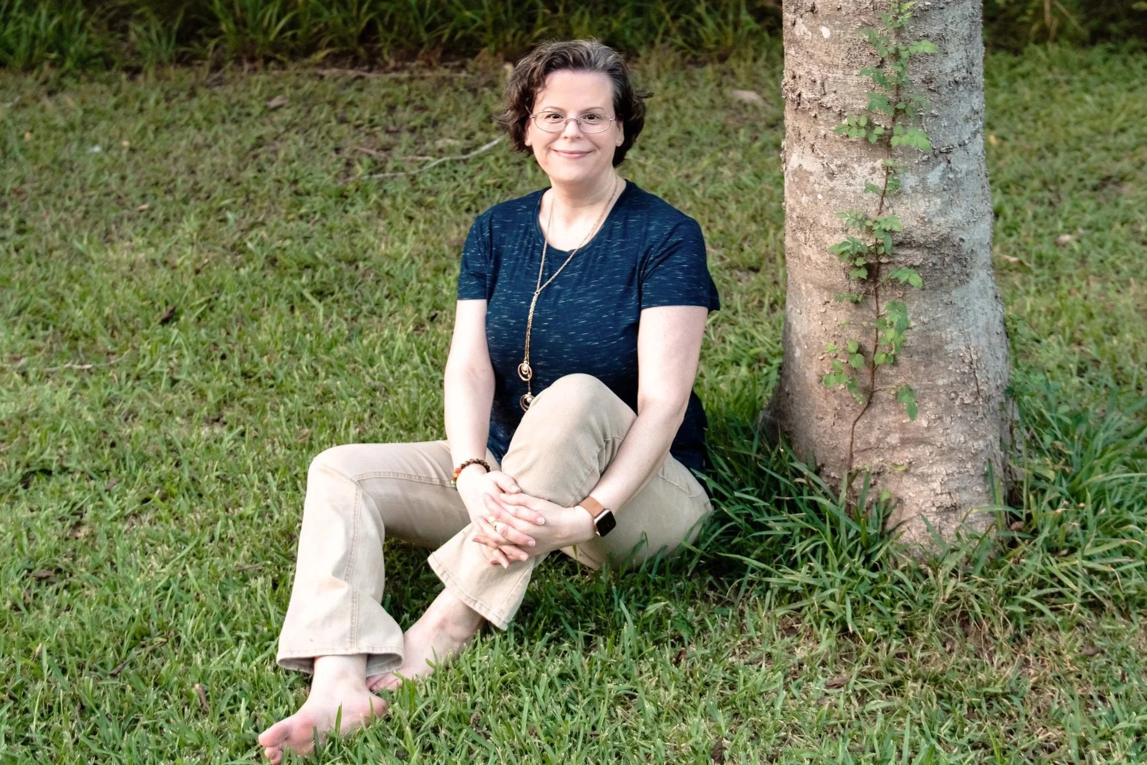Woman sitting on the grass with next to a tree without shoes.