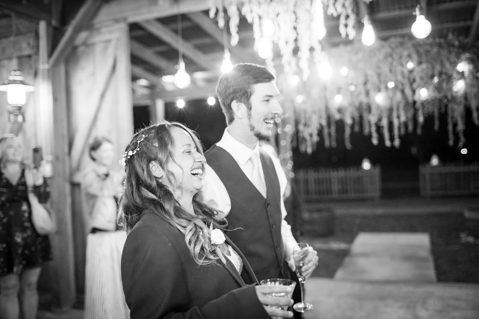 A black and white photo of a wedding reception with a smiling bride and groom holding drinks, surrounded by guests in a rustic venue decorated with string lights and hanging foliage.